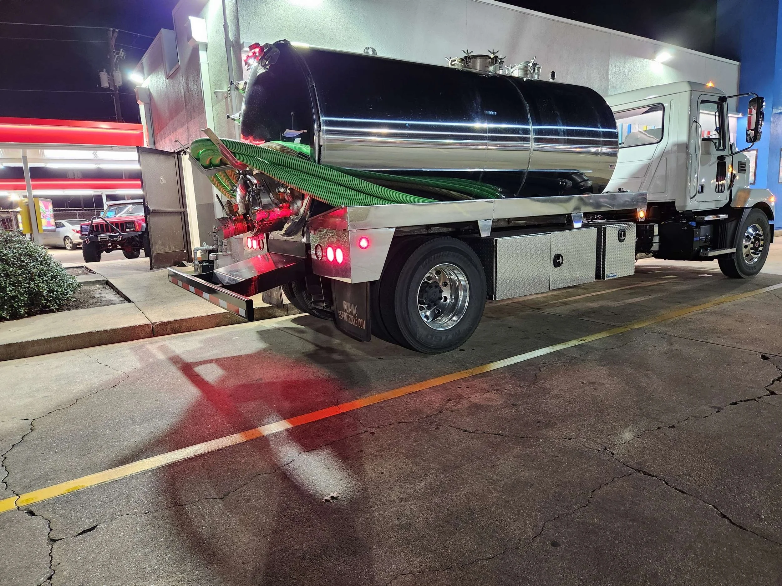 A large commercial water tanker truck parked outside a building at night, with hoses and equipment on its side, in a parking lot with cars and a convenience store in the background.