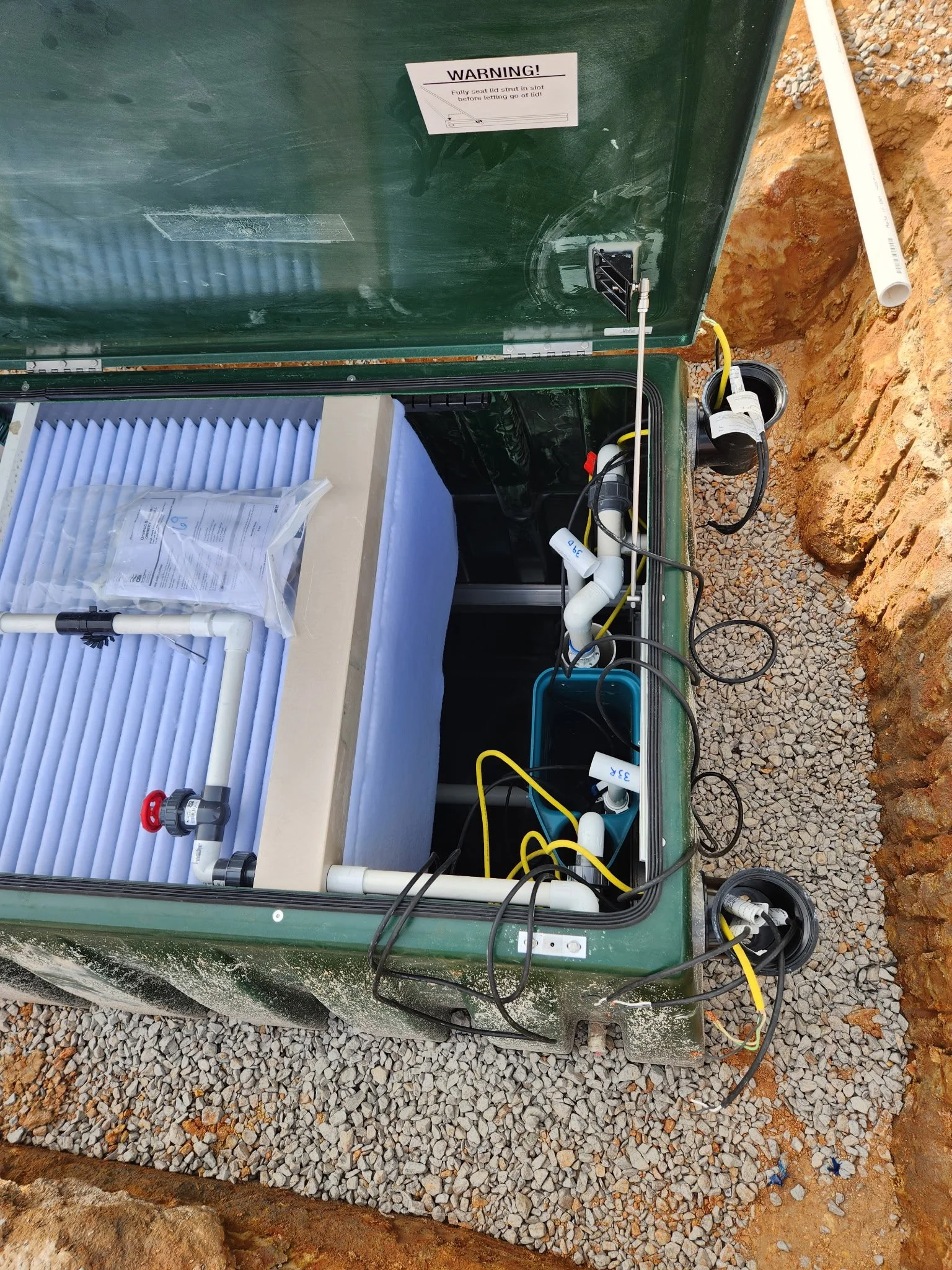 An outdoor water treatment or filtration system installed in a gravel trench, with pipes and electrical wiring connected, and a warning sign on the green metal cover.