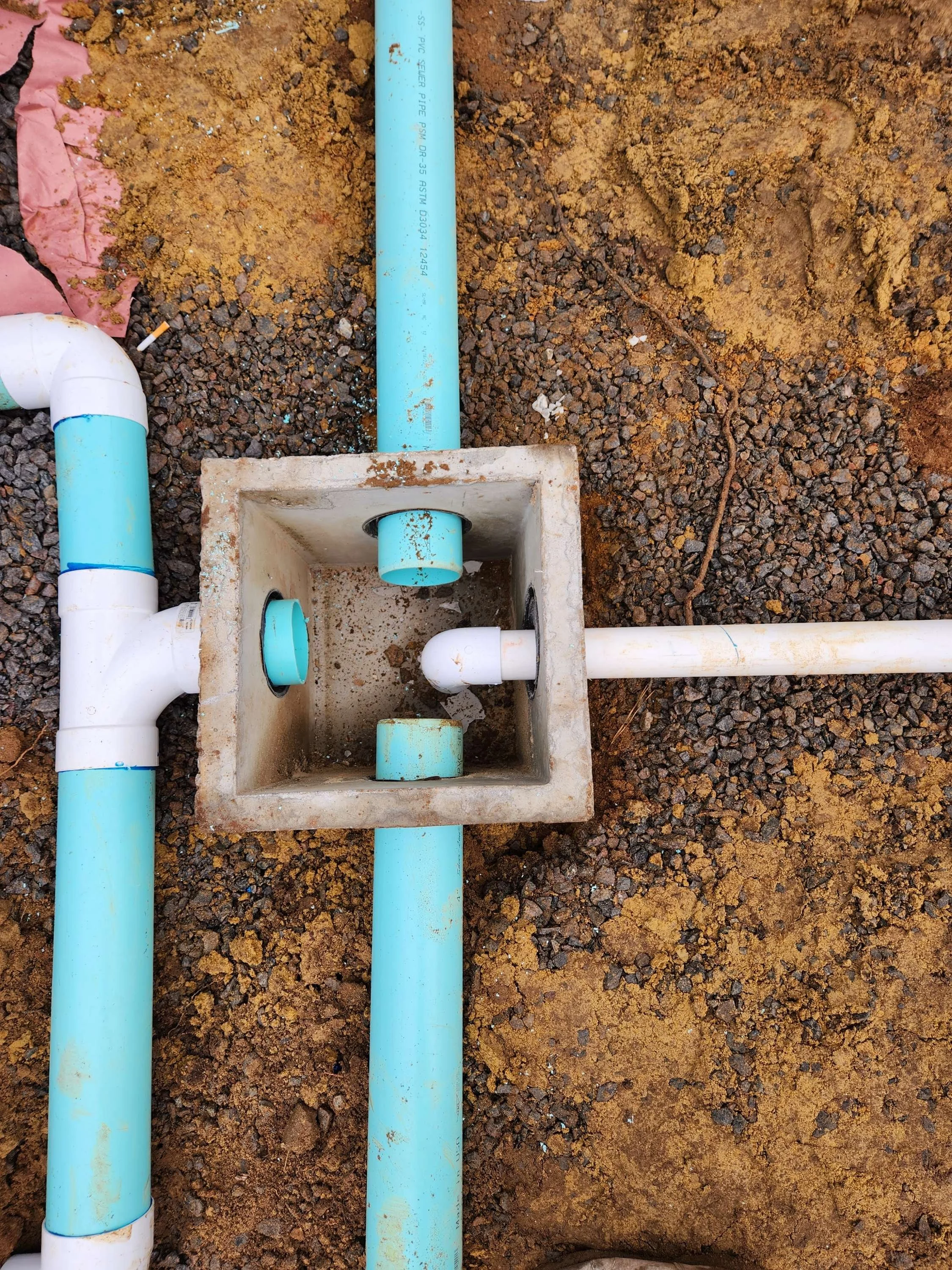 Inside view of an outdoor plumbing system with blue and white PVC pipes connected to a concrete box, on gravel ground with soil nearby.