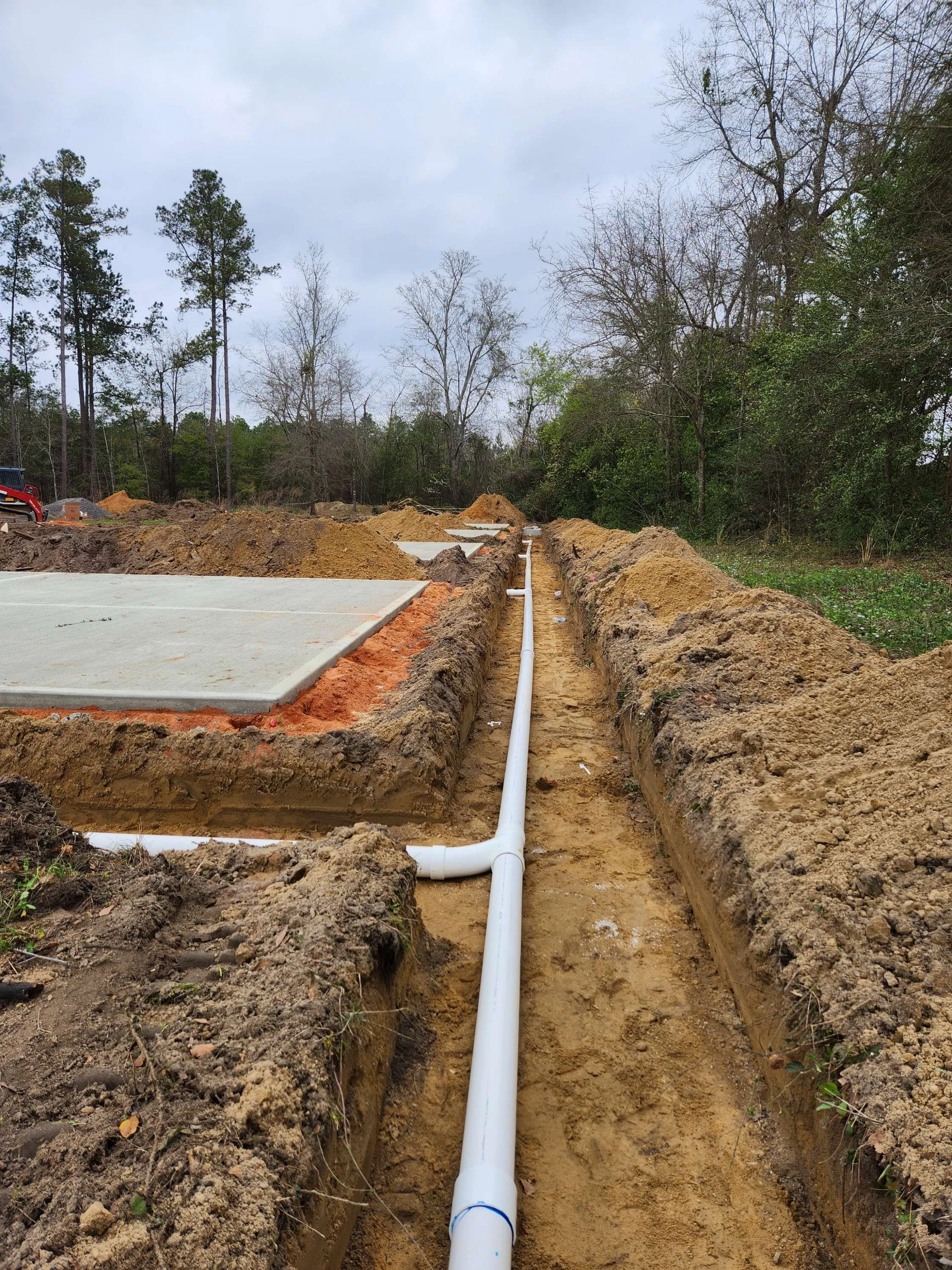 Construction site with concrete slabs and a white pipe running through a trench, surrounded by dirt and trees in the background.