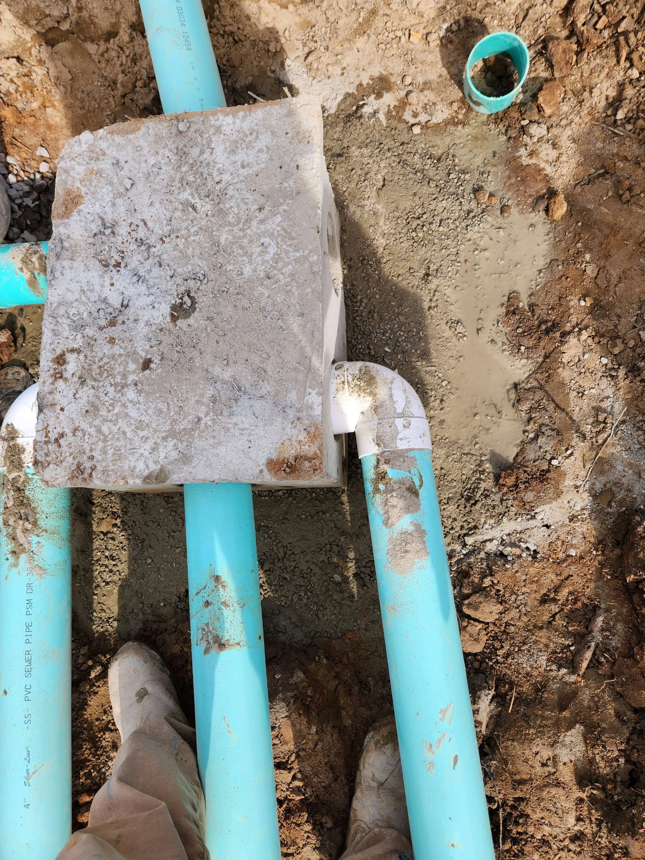 Construction site view from above showing blue PVC pipes, a concrete cover, dirt, and a small pipe protruding from the ground.
