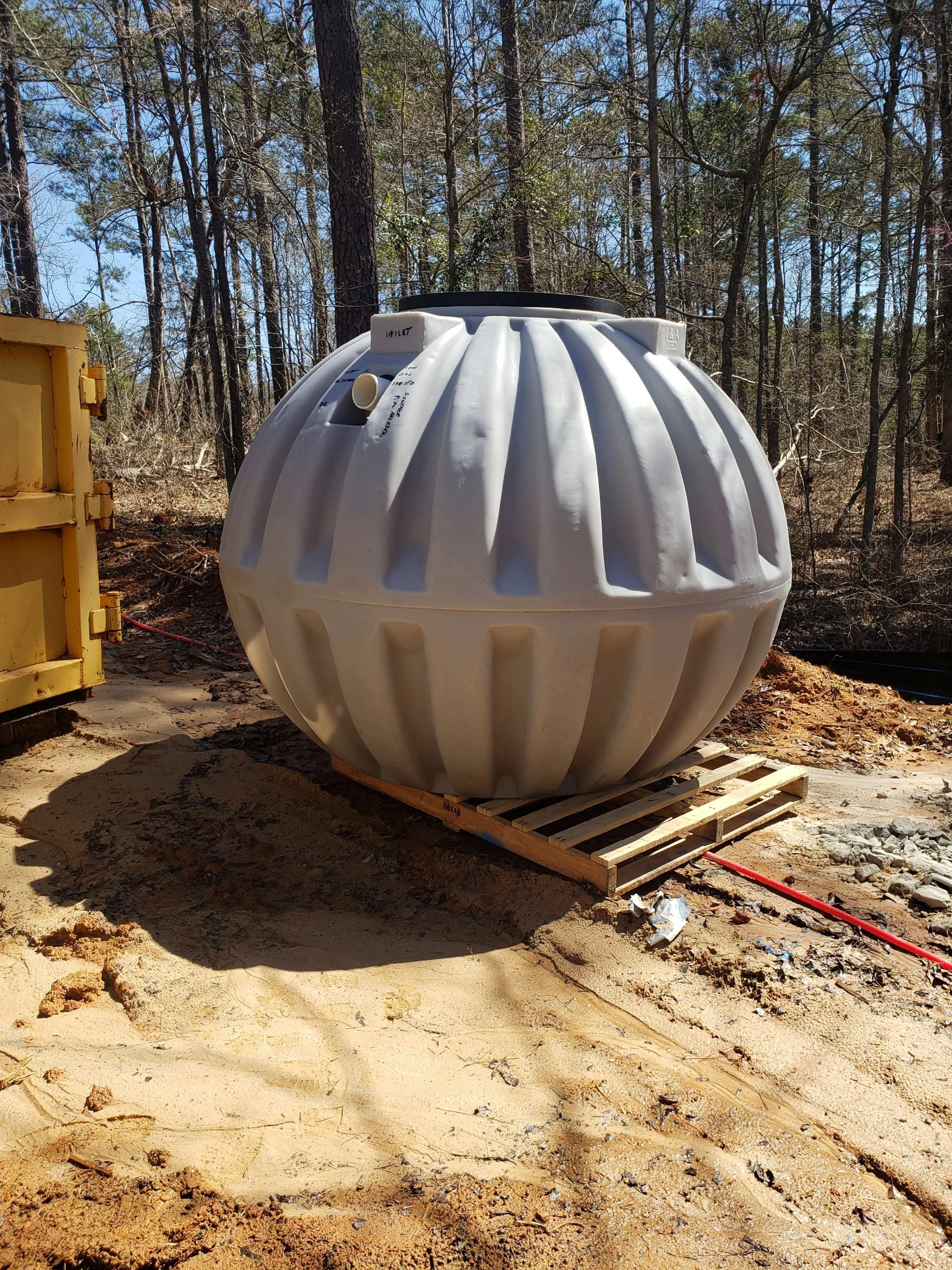 A large, round, gray plastic tank with vertical ridges, placed on a wooden pallet at a construction site surrounded by trees and dirt.