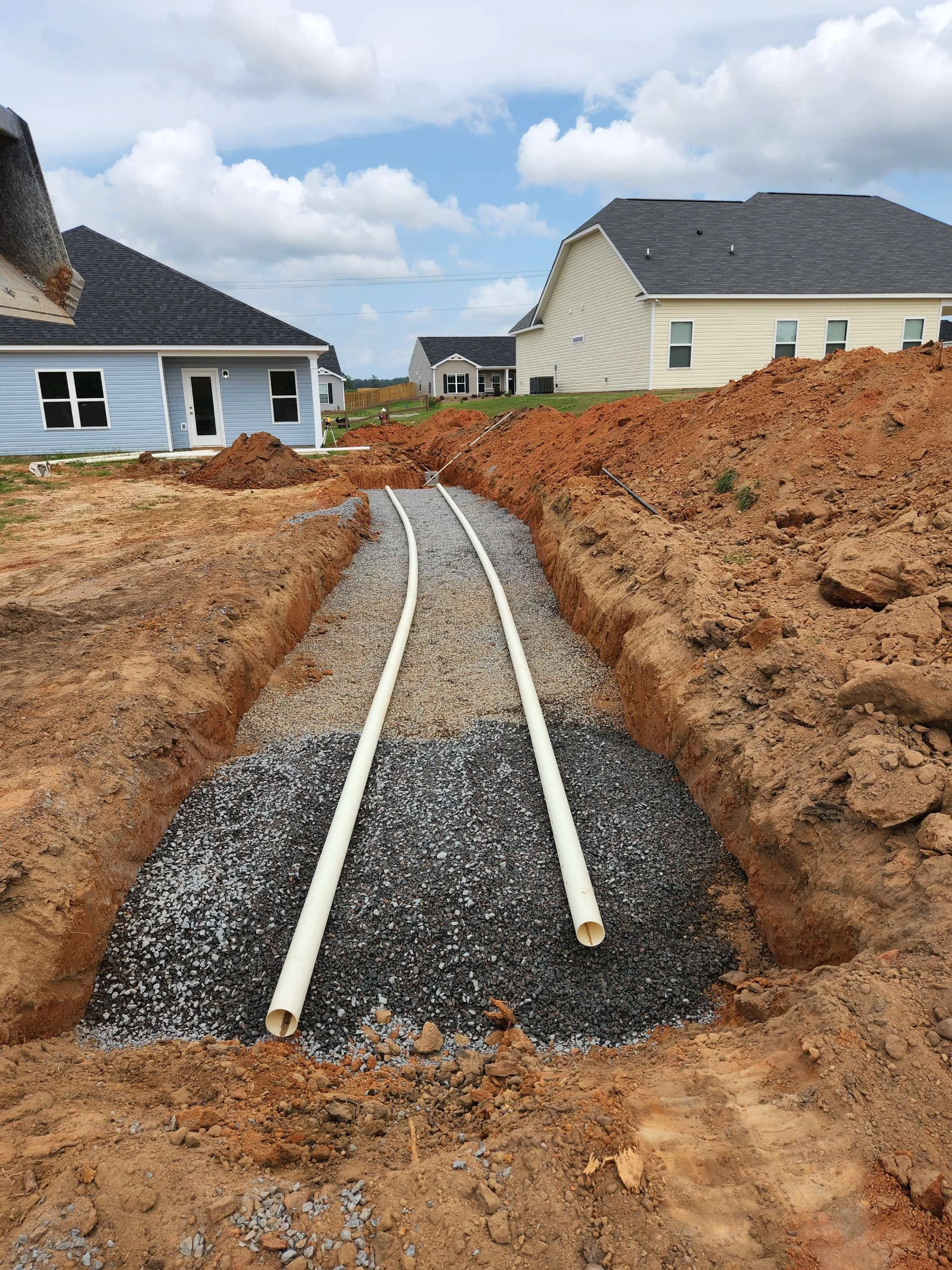 Construction site with two white pipes being laid in a gravel trench surrounded by dirt on both sides, in a residential neighborhood with houses and partly cloudy sky in the background.