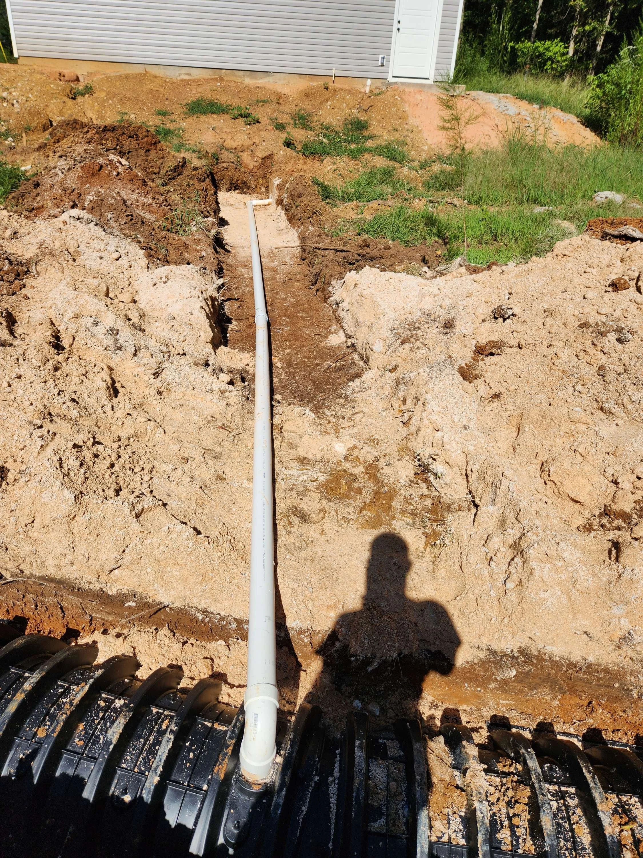 Underground water piping installation at a construction site with a large trench, white PVC pipe, and a drainage or utility cover in the foreground.
