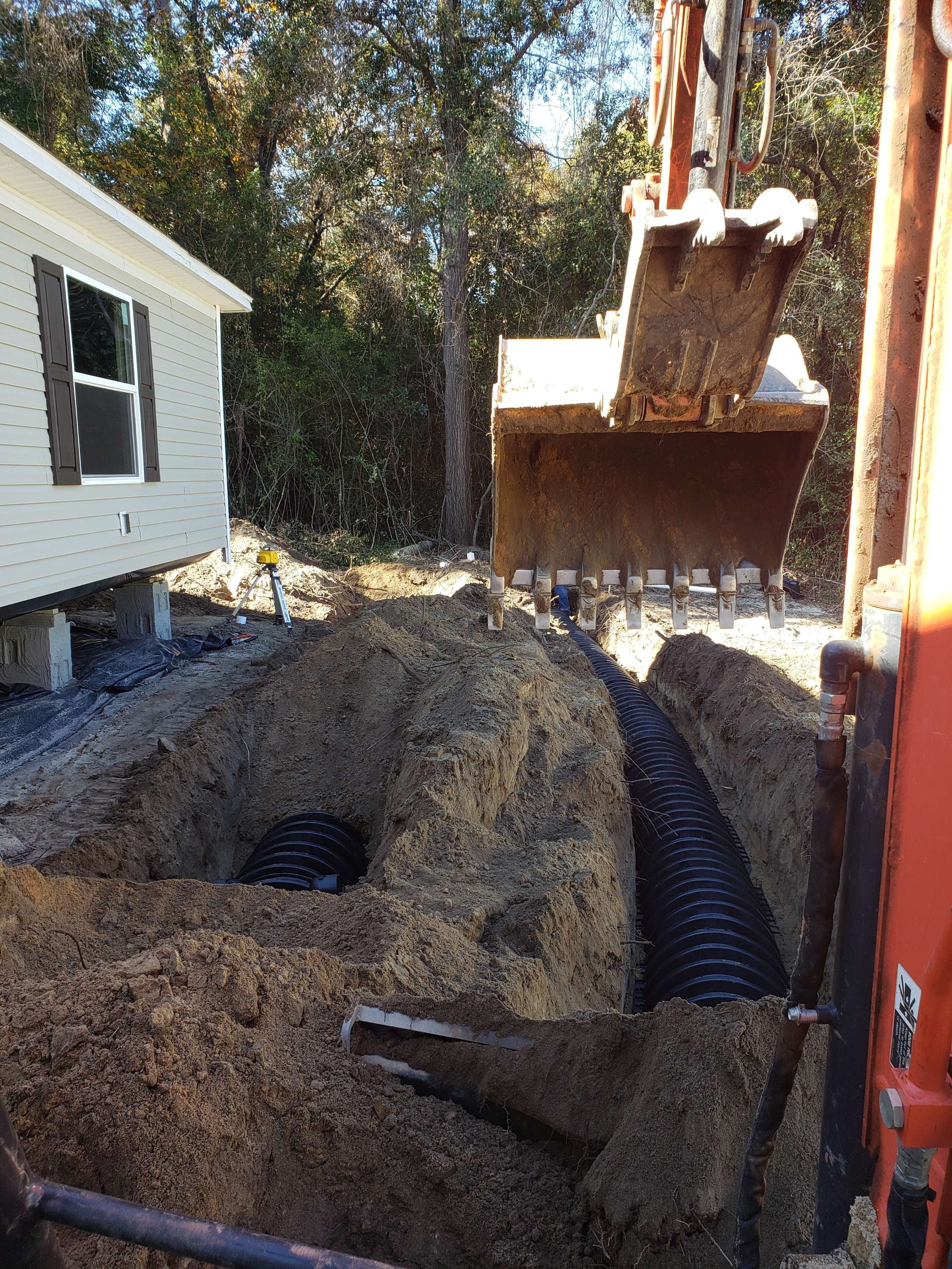 Construction site with a trench and black corrugated pipes, excavator bucket hanging overhead, house on the left, and wooded area in the background.