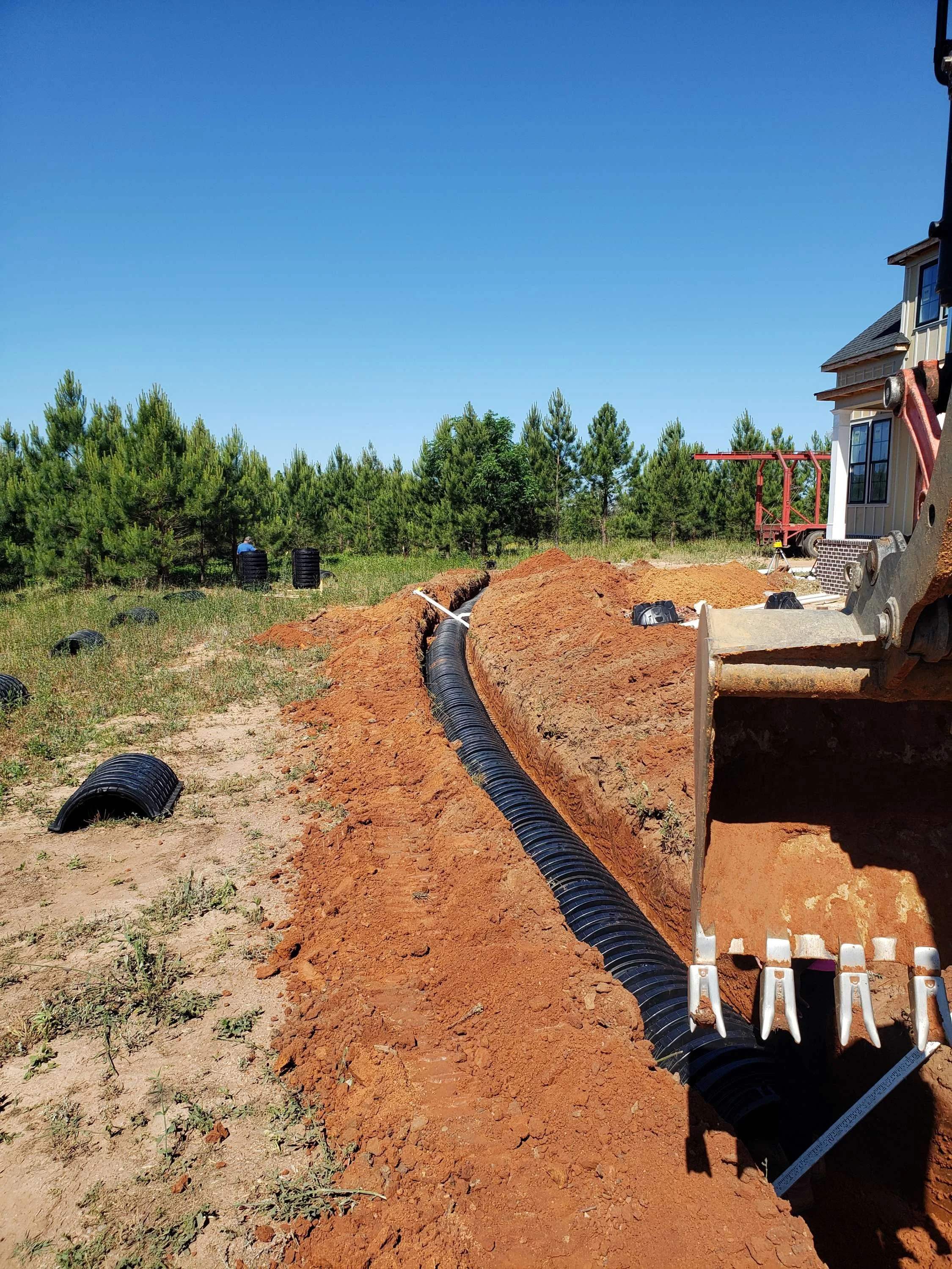 Construction site showing a trench with a black corrugated pipe being installed, surrounded by dirt and some black circular objects on the ground, with trees in the background and part of a house on the right.