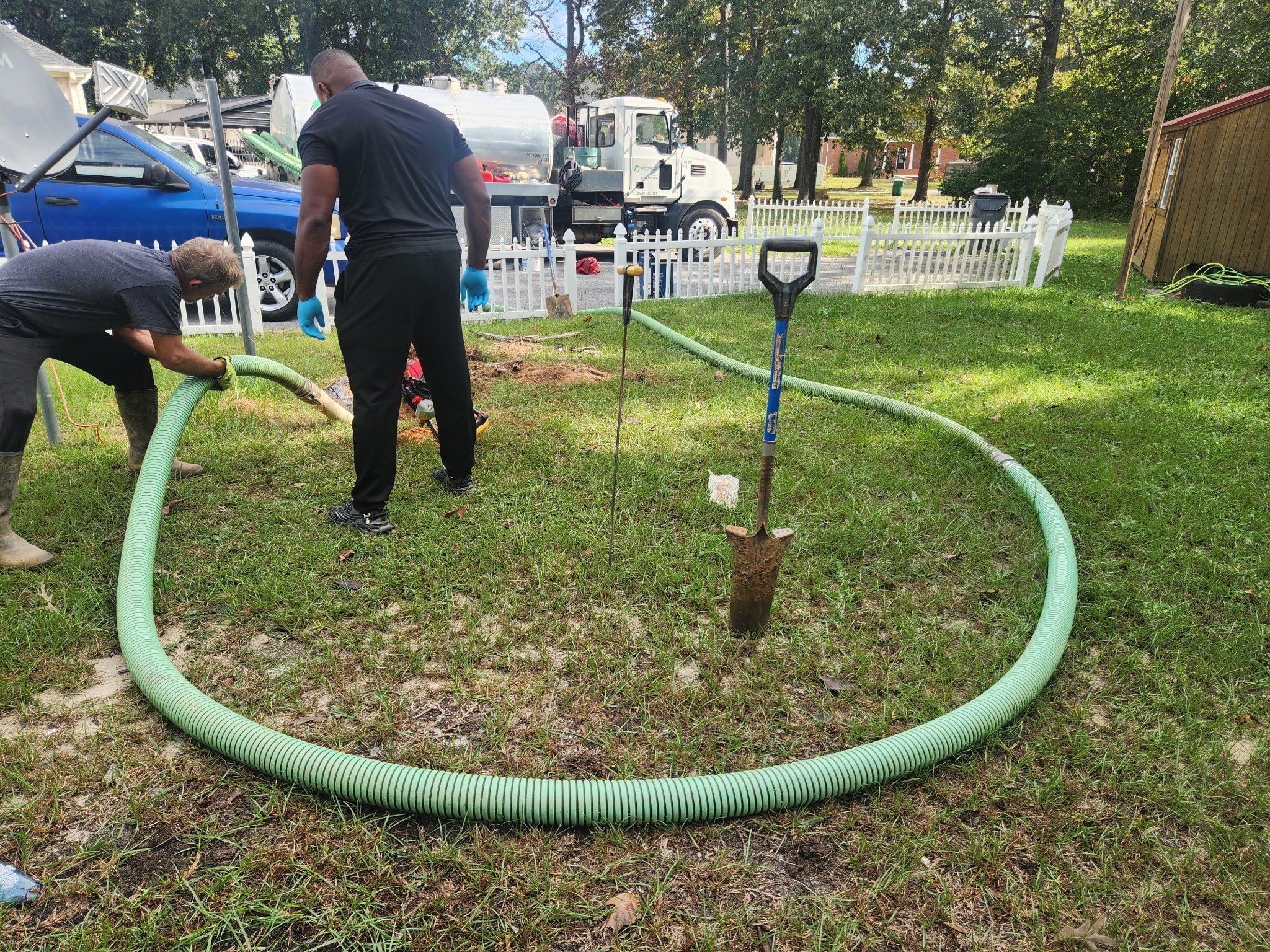 Two workers install a large green flexible pipe in a yard, with shovels and construction tools nearby.