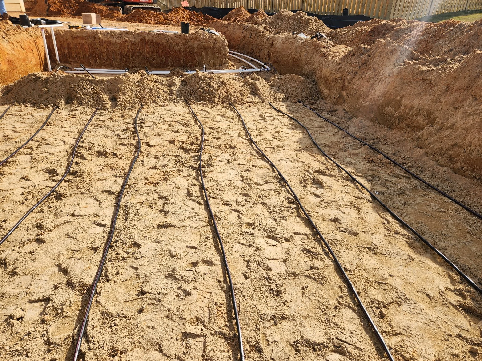 Construction site with irrigation tubing laid out on sandy ground, surrounded by dirt mounds and excavation trenches.