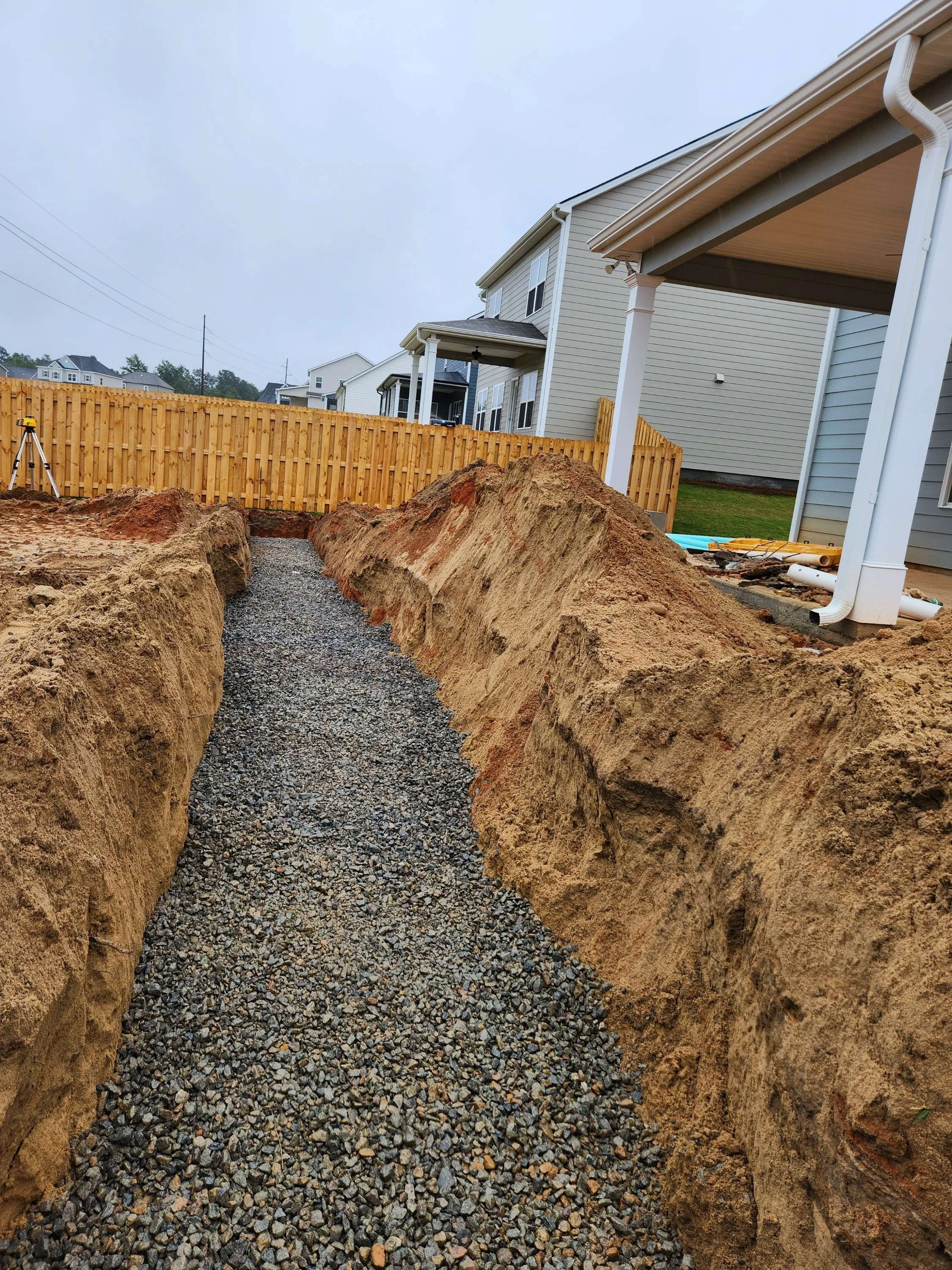 A construction site with a dirt trench filled with gravel, surrounded by large dirt mounds, in a residential backyard with houses, a wooden fence, and a partly cloudy sky.