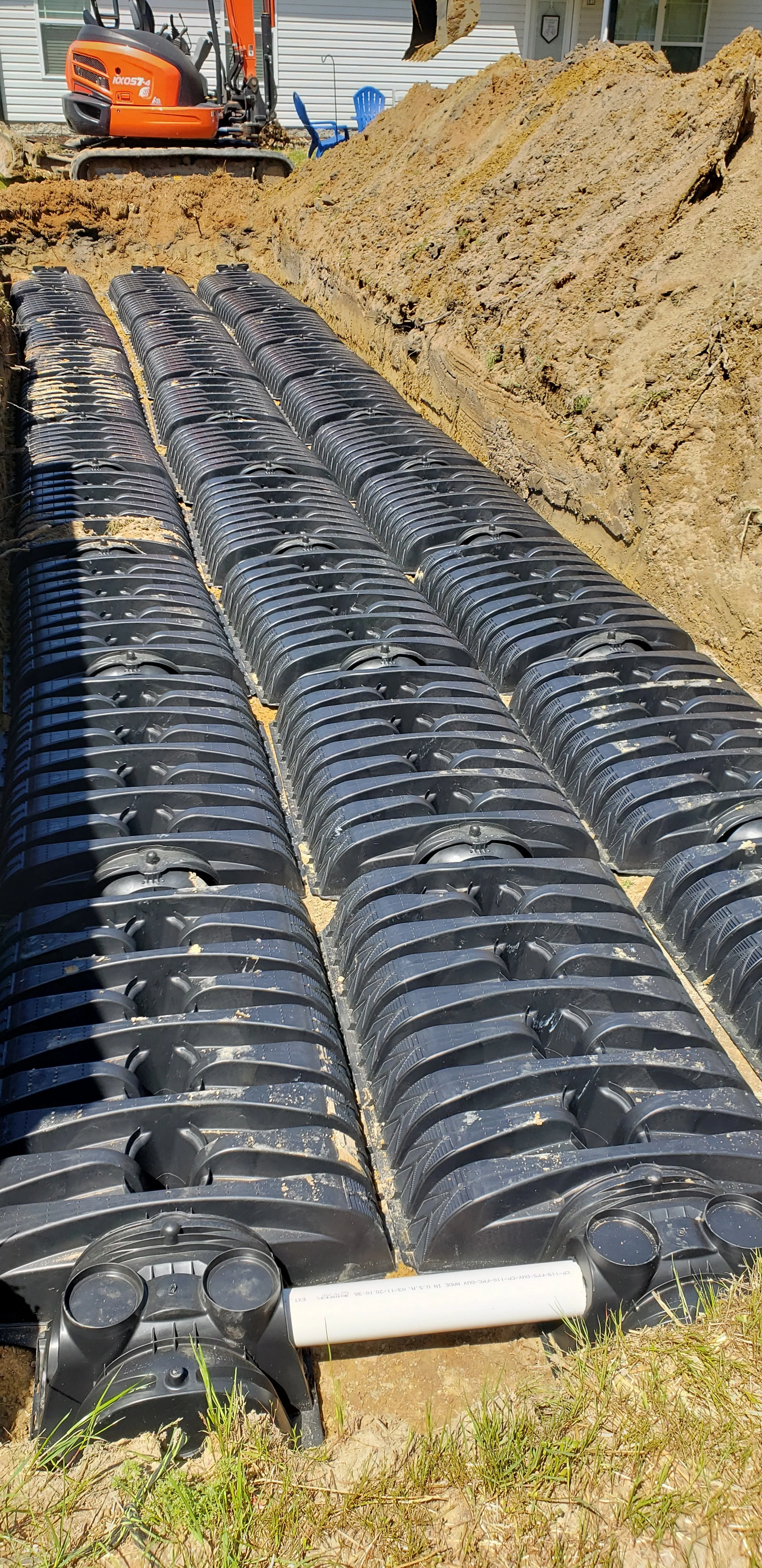 Multiple black plastic drainage drainage chambers installed in a trench with an orange excavator in the background.