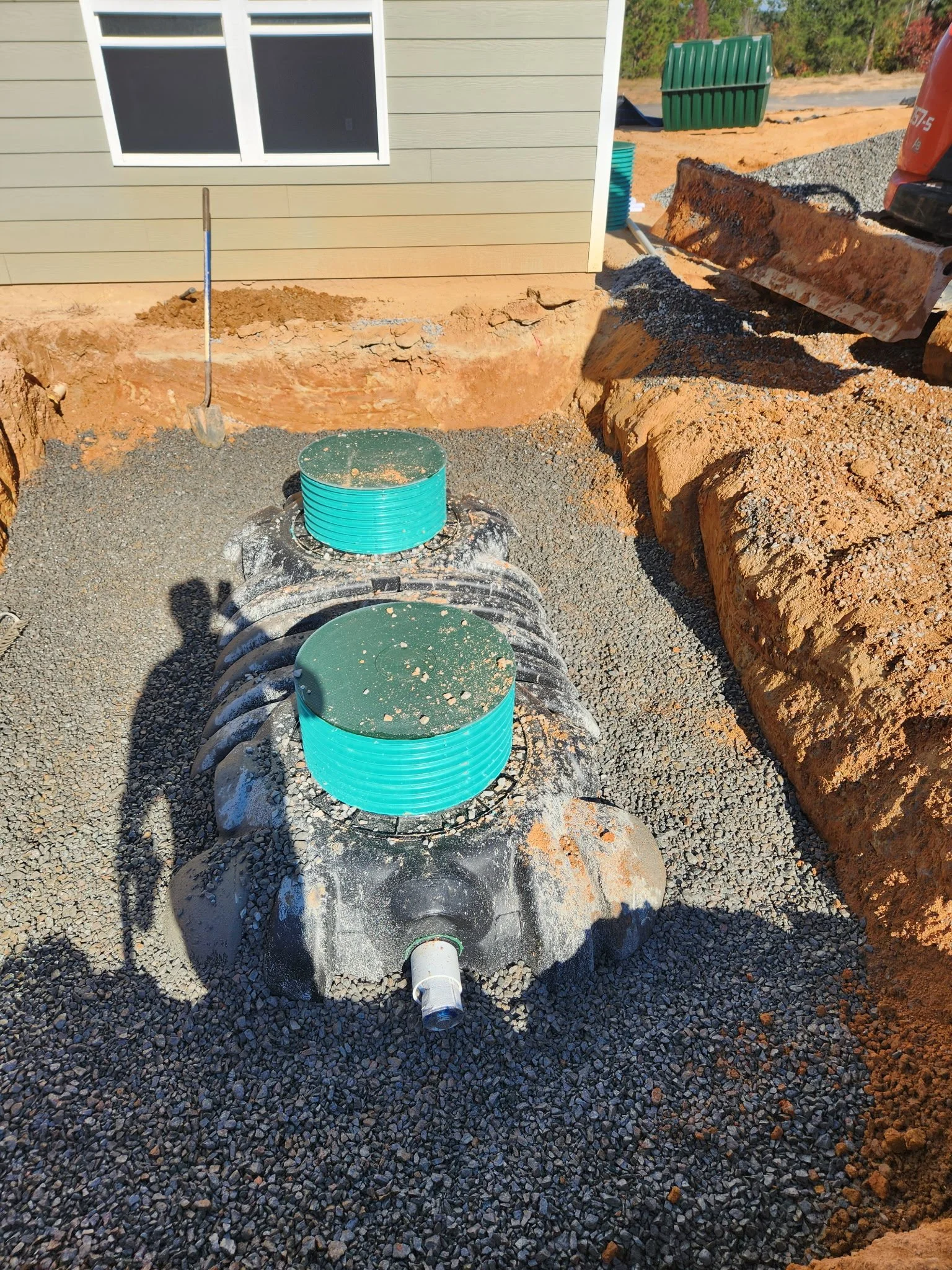 Construction site showing a residential yard with gravel. There is a septic tank with two access ports on top and a pipe at the front. A shovel and an excavator are also visible.
