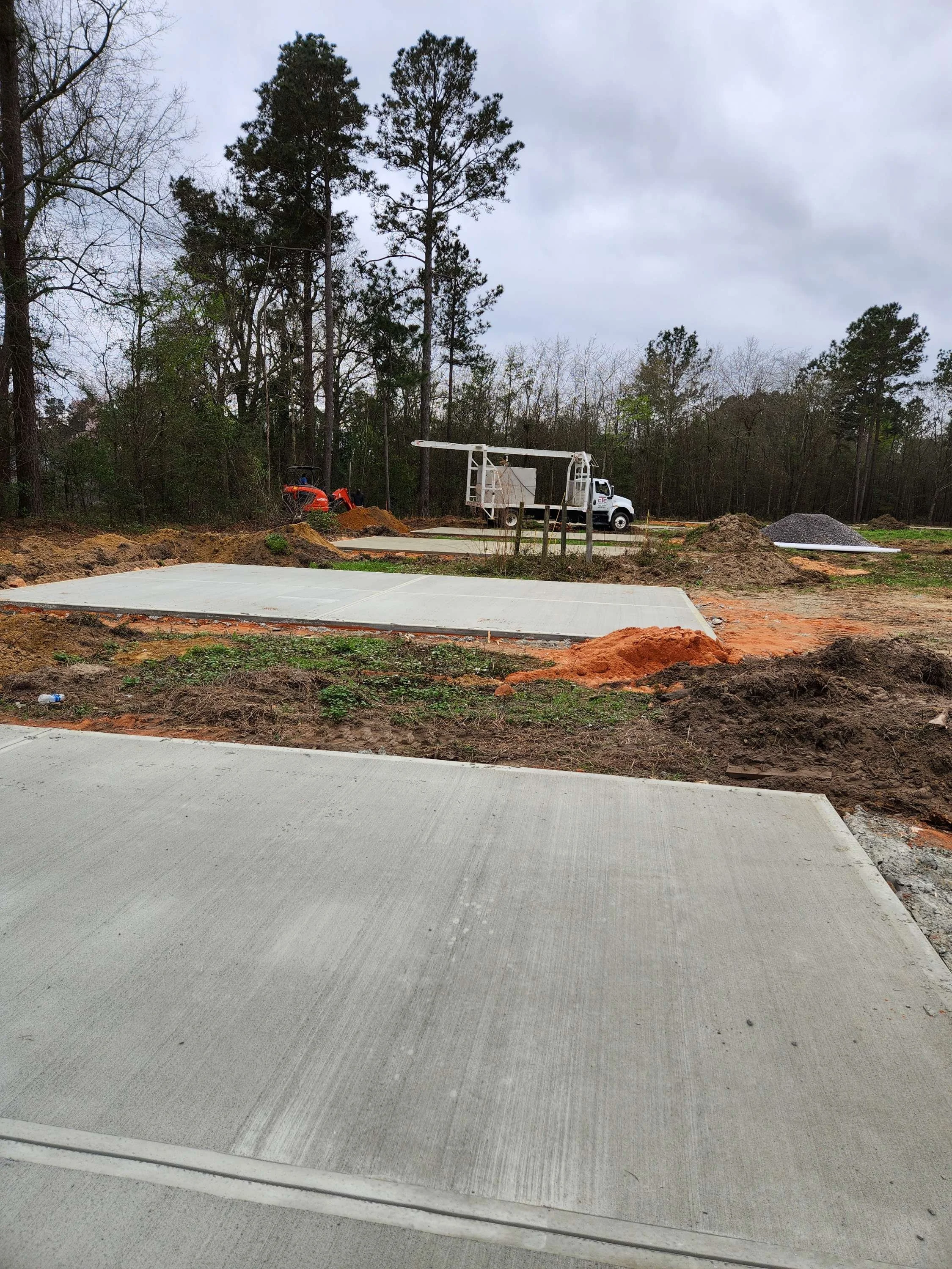 Construction site with freshly poured concrete slabs, a dump truck, and construction equipment in a wooded area.