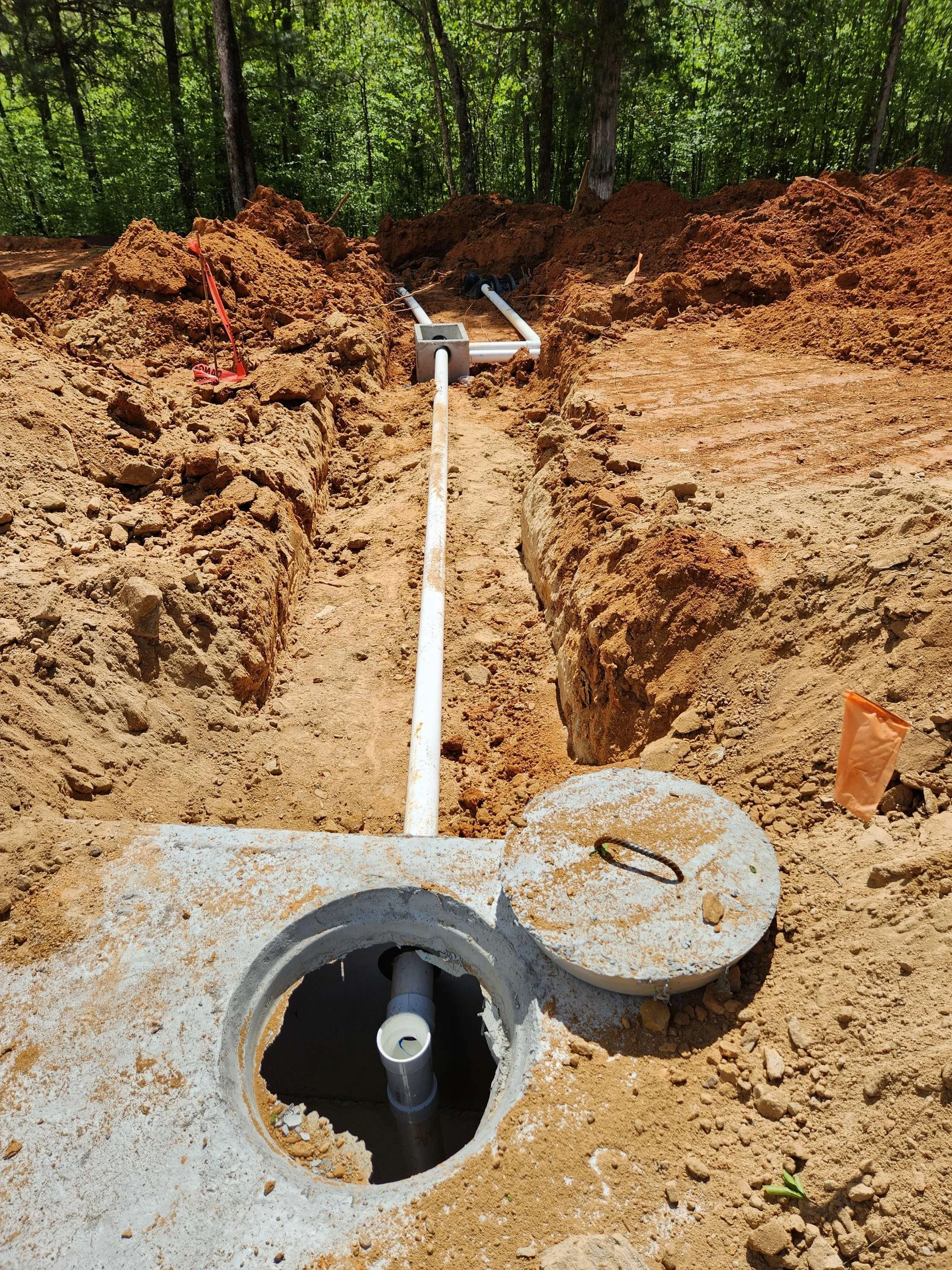 Underground utility installation in progress with pipes and conduit in a trench between dirt mounds in a wooded area.