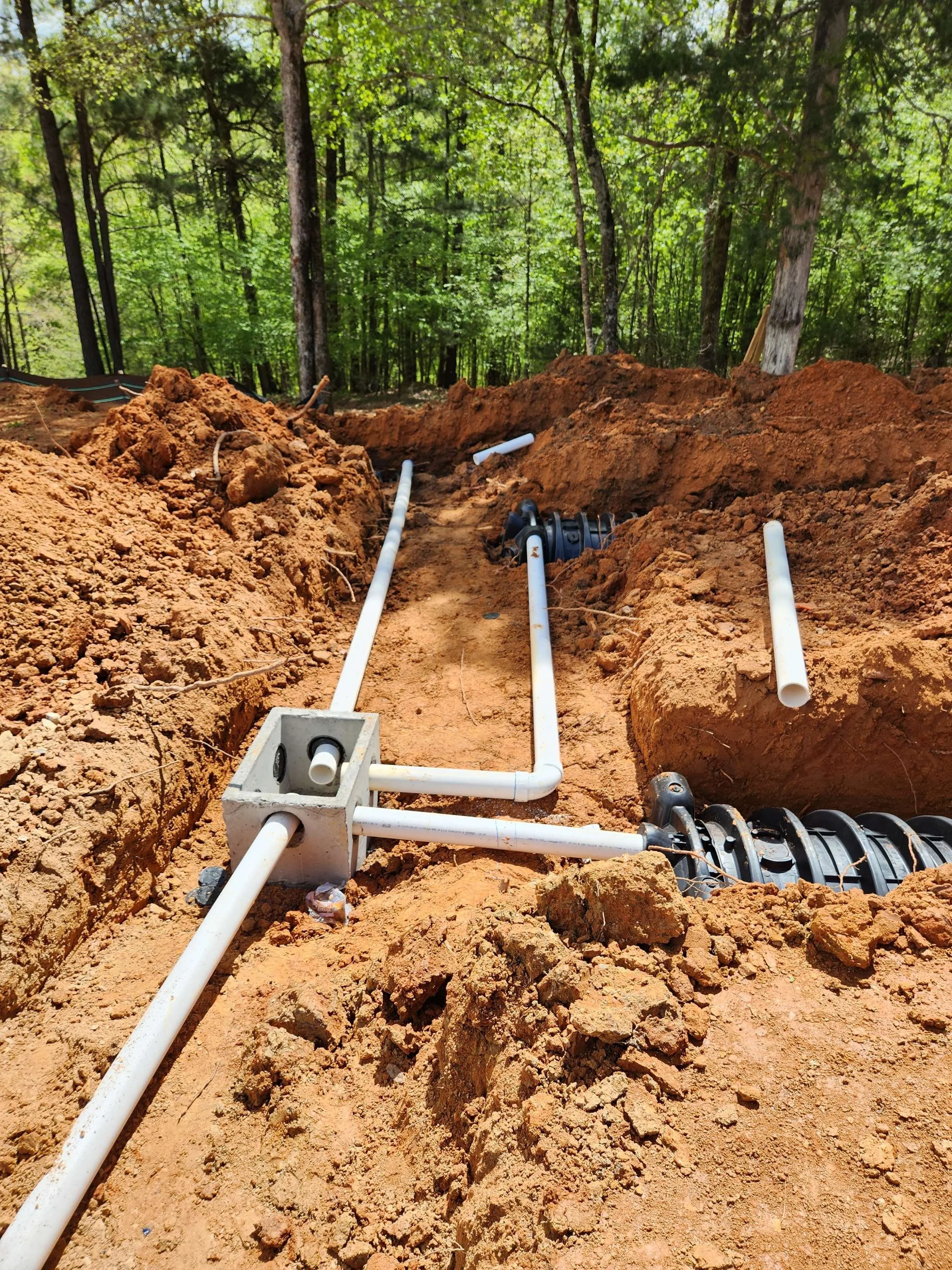 Construction site with plumbing pipes laid in an excavated trench surrounded by soil and trees.