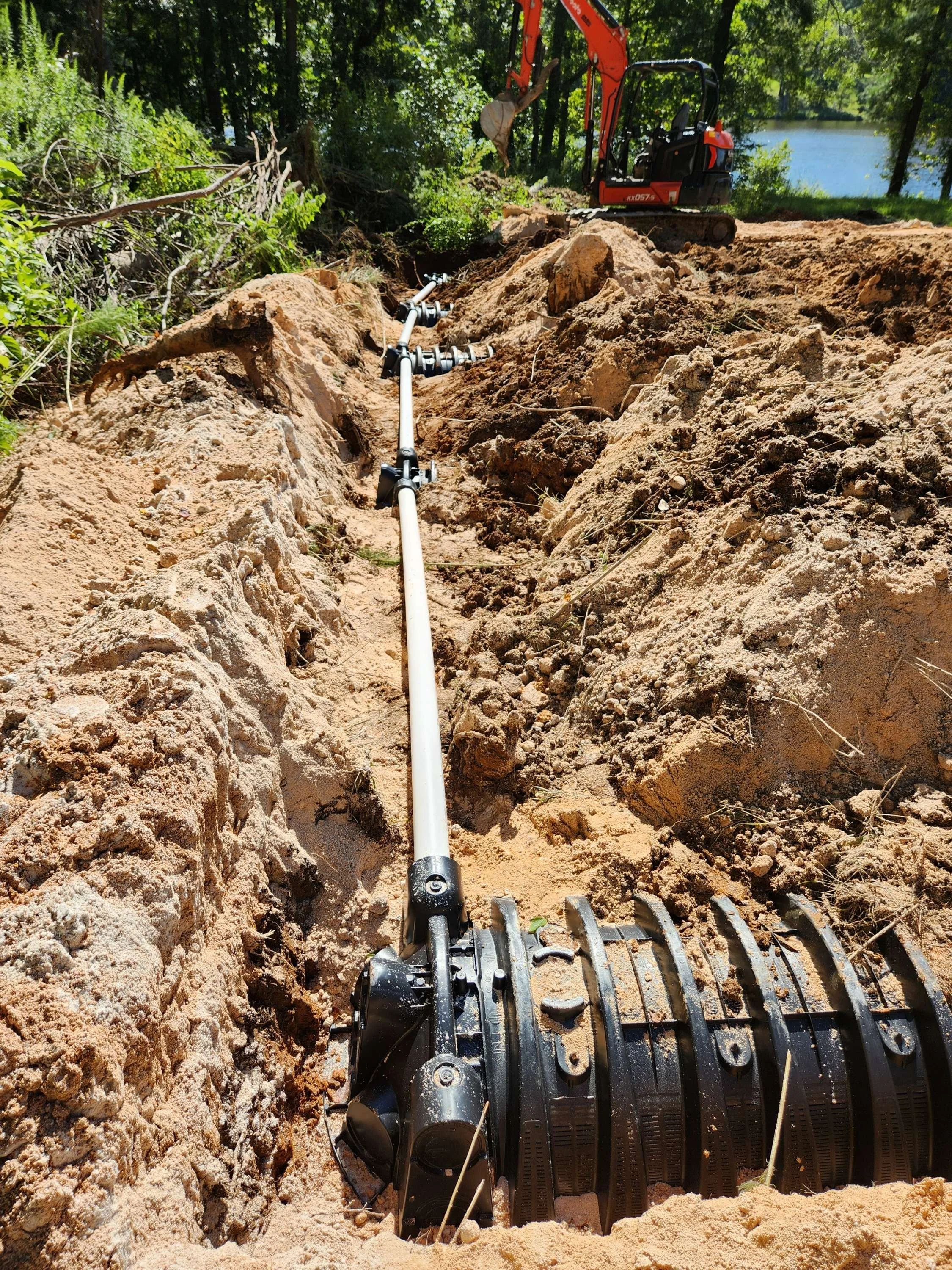 Underground pipeline installation with a small excavator and new pipe underground, near a lake surrounded by trees.