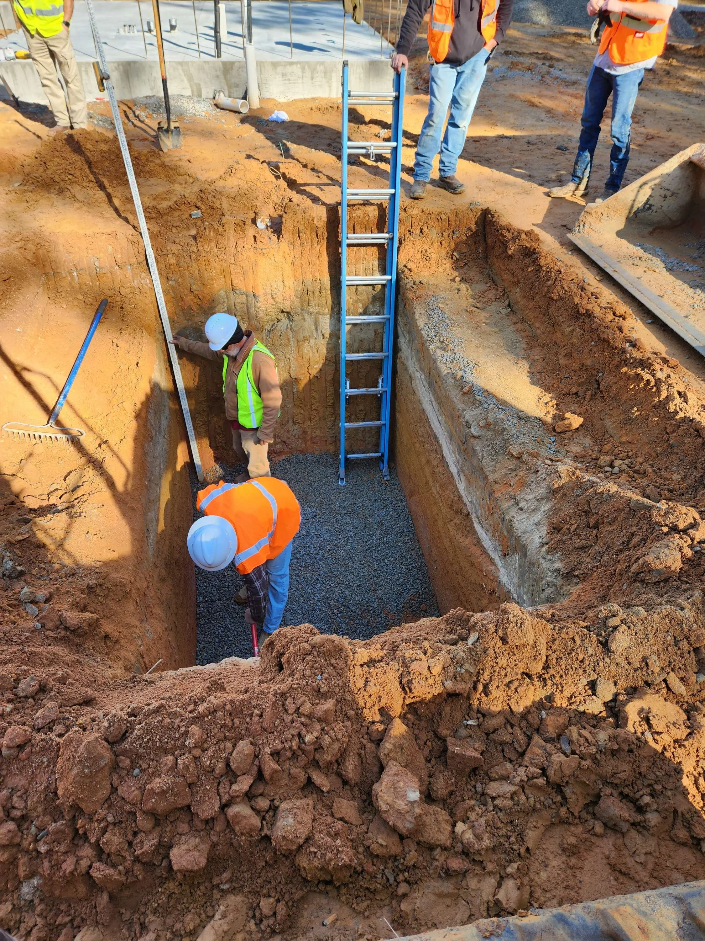 Construction workers in safety vests and hard hats working in a deep excavation, with a ladder and equipment on a construction site.