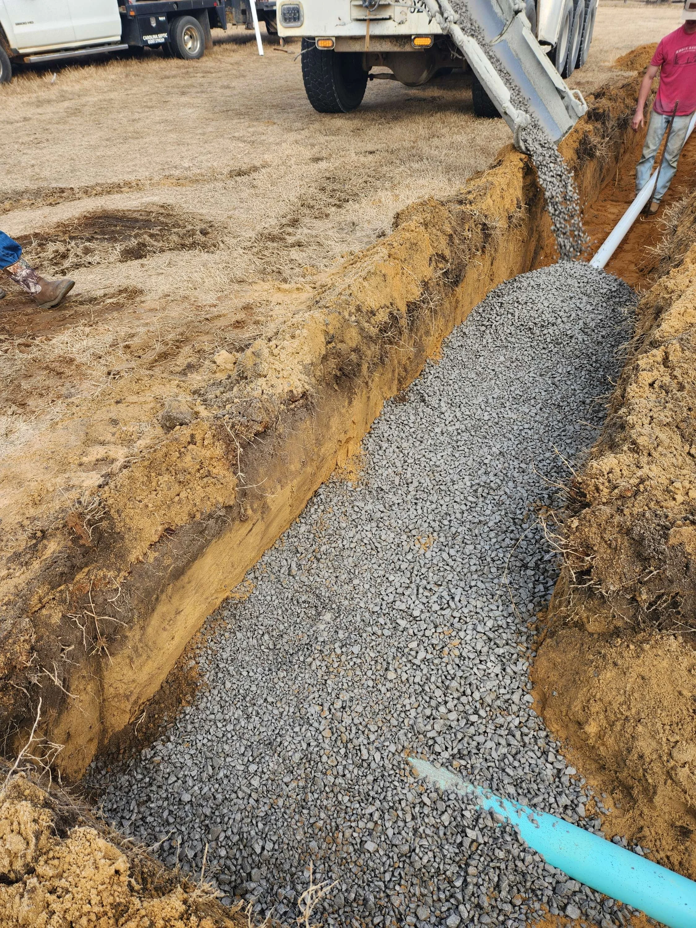 A construction site with a trench filled with gravel, a worker in a pink shirt holding a shovel, and a large truck pouring gravel into the trench.