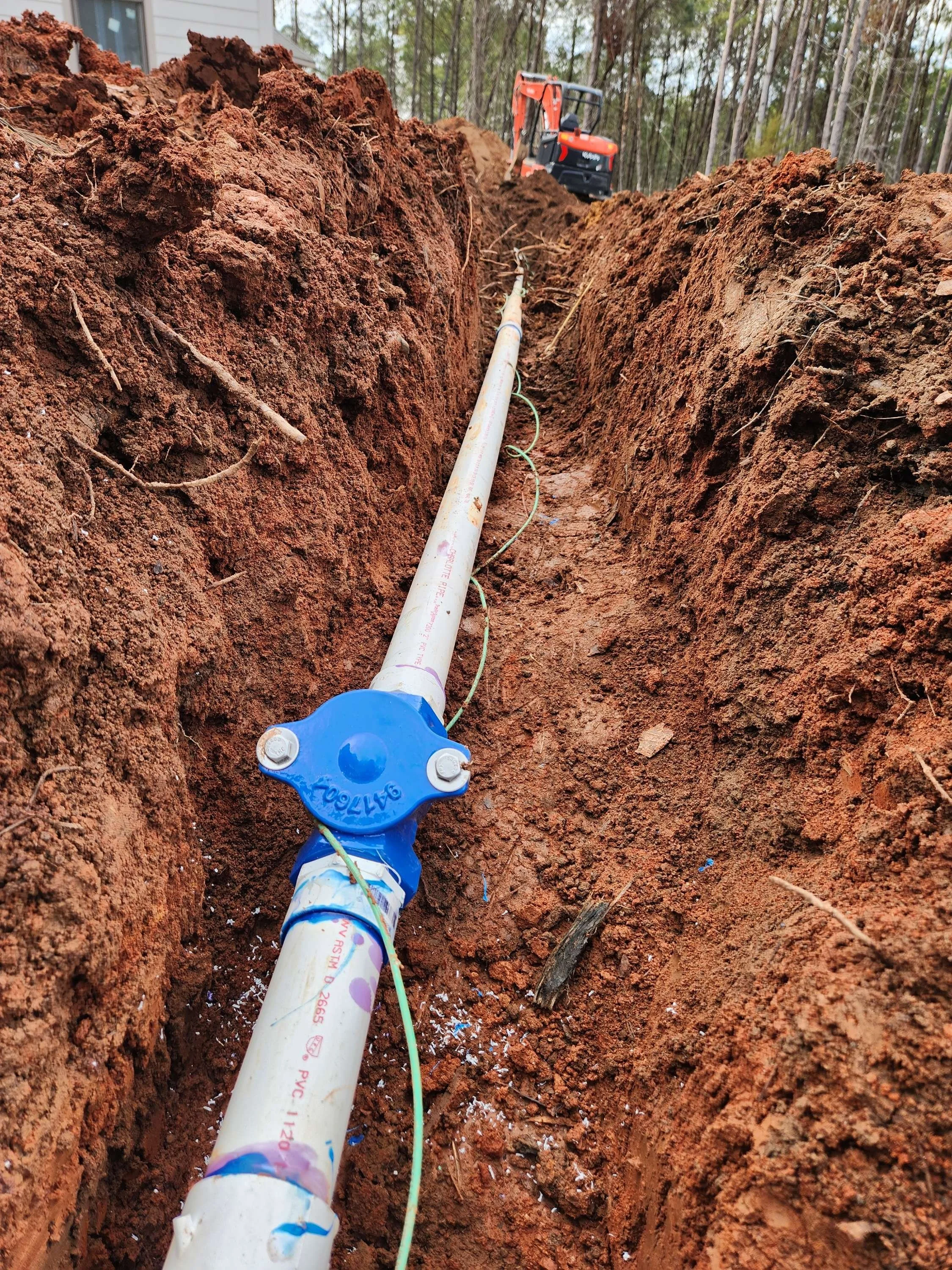 A trench with a white pipe laid inside and connected to a blue valve, with a construction site and forest in the background.