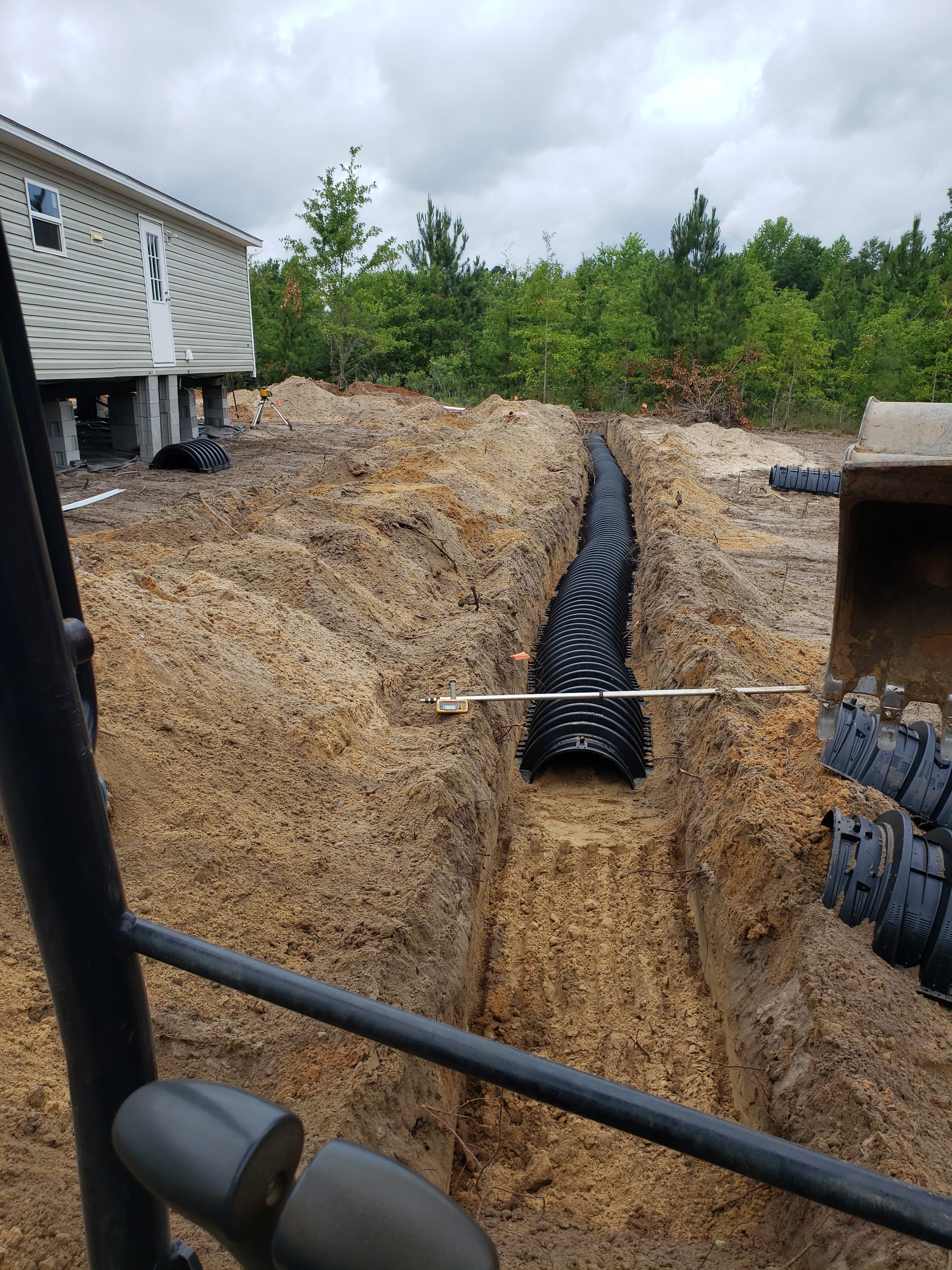 Construction site showing a trench with black corrugated piping, soil mounded on both sides, and trees in the background.