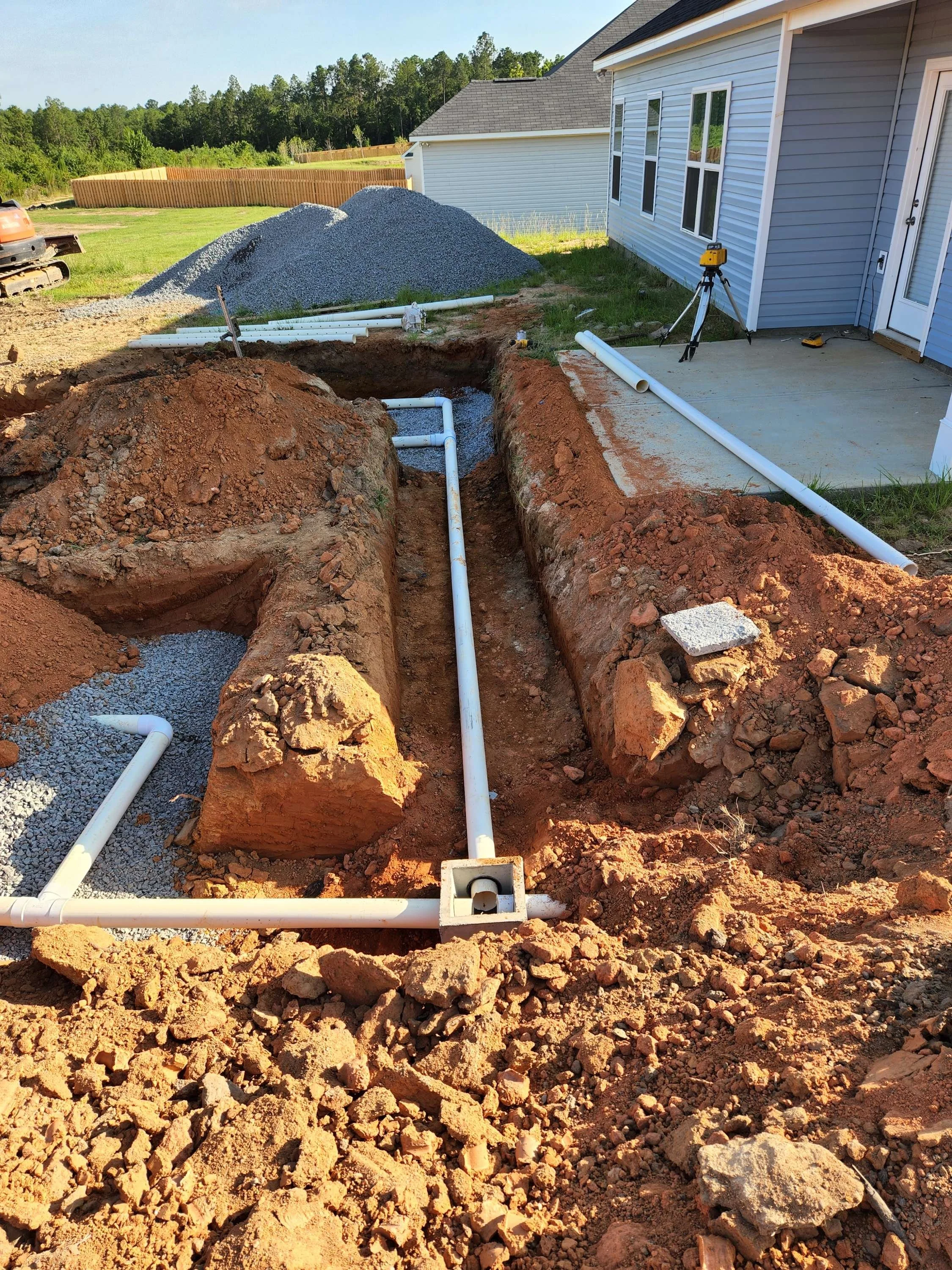 Underground plumbing installation with PVC pipes in a backyard trench next to a house, with construction equipment and gravel pile in the background.