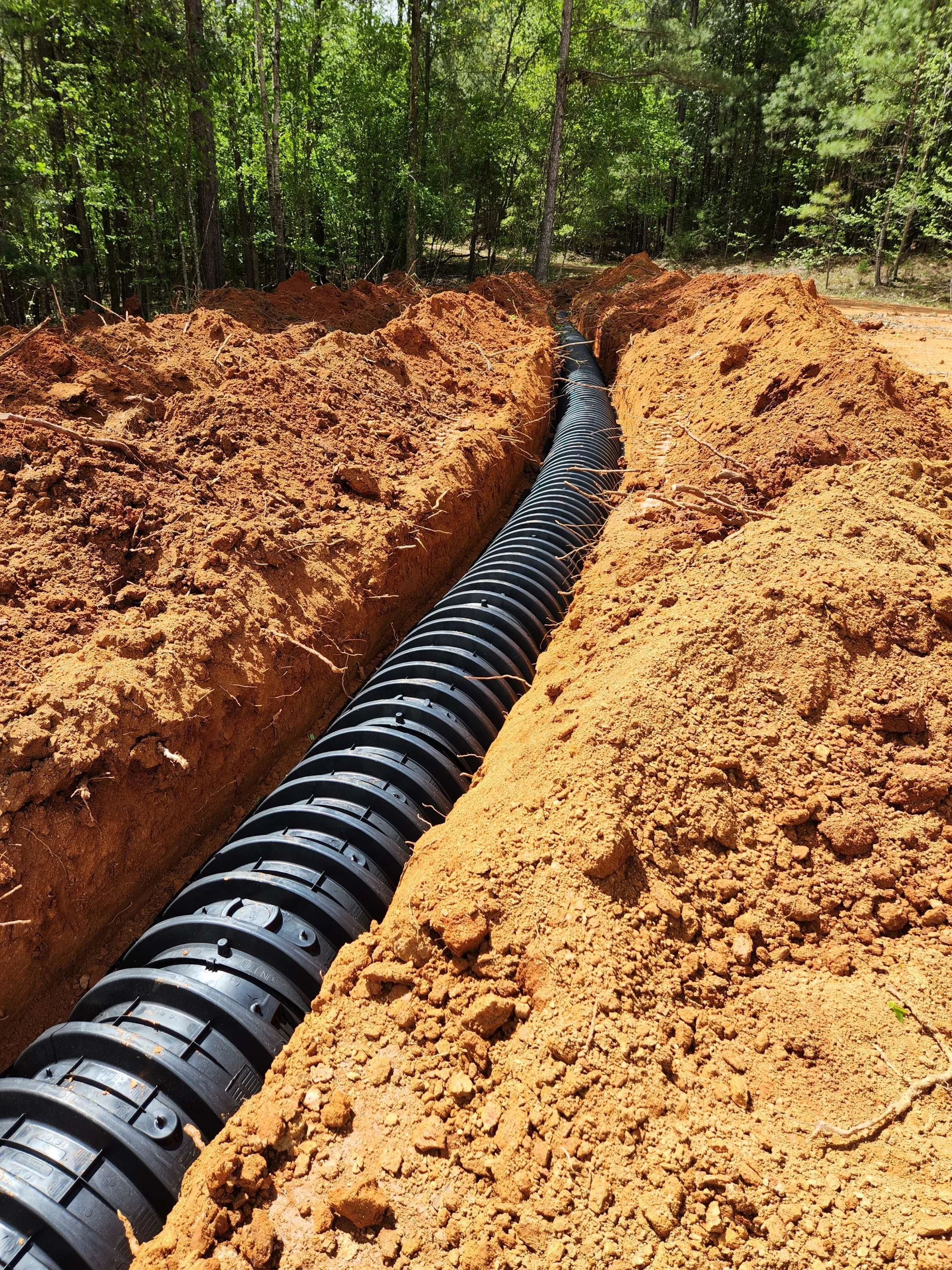 A black drainage pipe runs through a trench in the ground, surrounded by dirt, with a forest of green trees in the background.