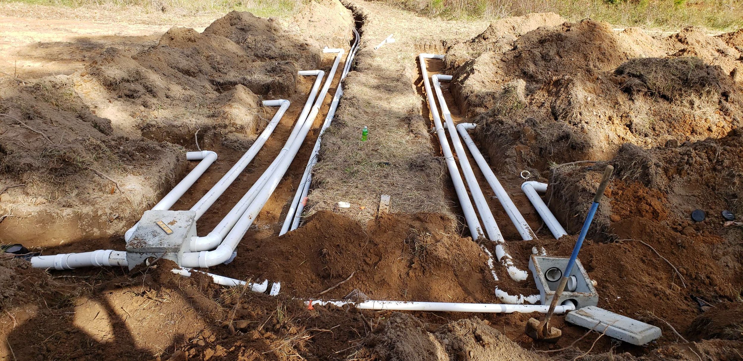 Underground utility pipes installation with several white pipes running through trenches in the dirt, with some pipes connected to junction boxes, and construction tools and materials visible around the site.