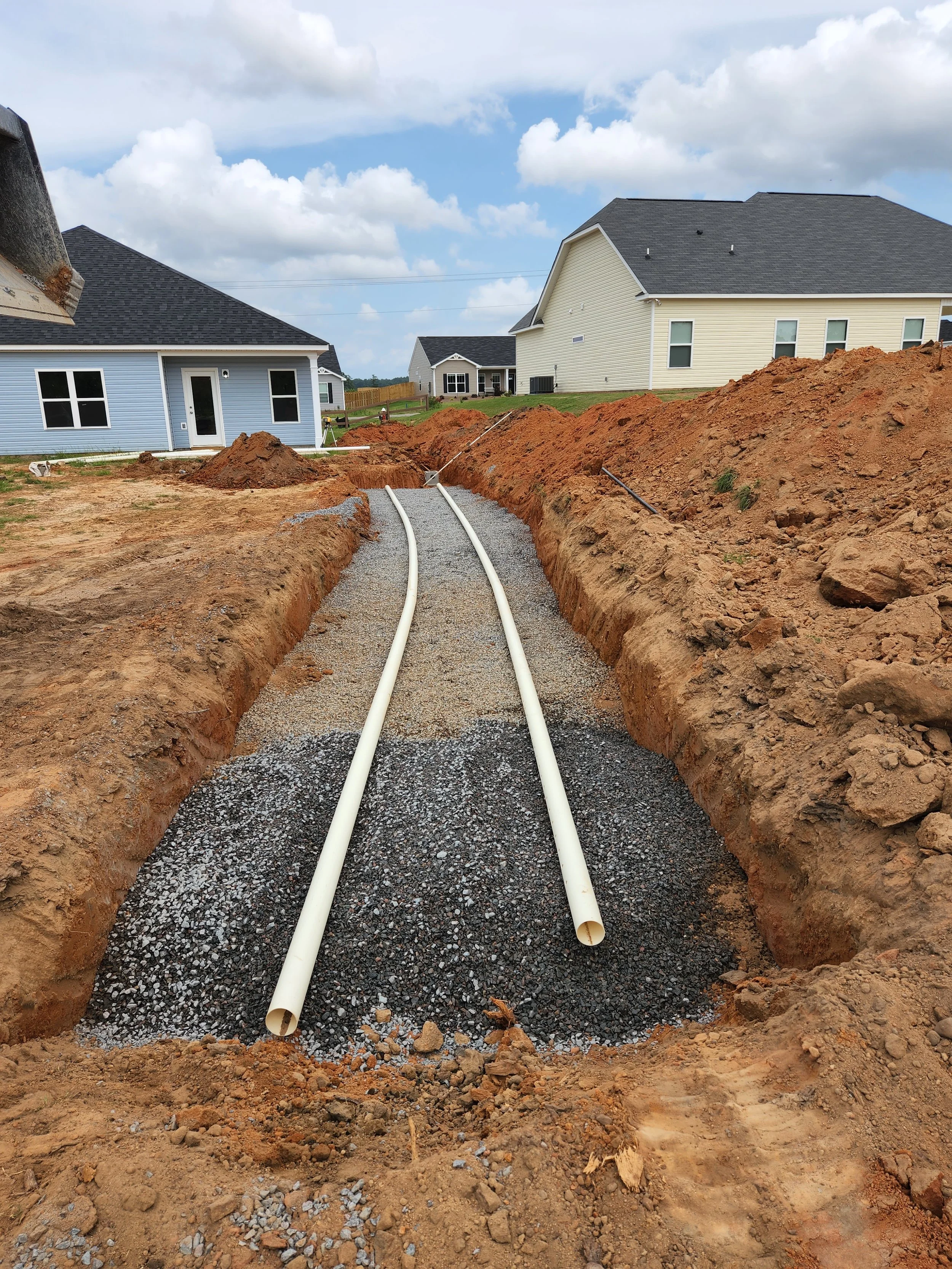 Construction site with two white pipes laid in a trench on a gravel base, surrounded by mounds of dirt and residential houses in the background under a partly cloudy sky.