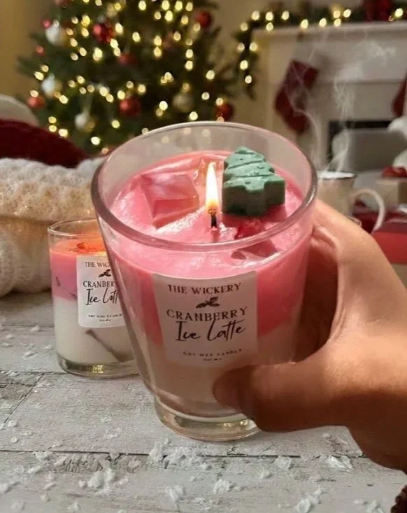 A person holding a pink candle with a small green decoration on top, labeled "Cranberry Ice Latte" from The Wickery, against a background of a decorated Christmas tree and festive decorations.