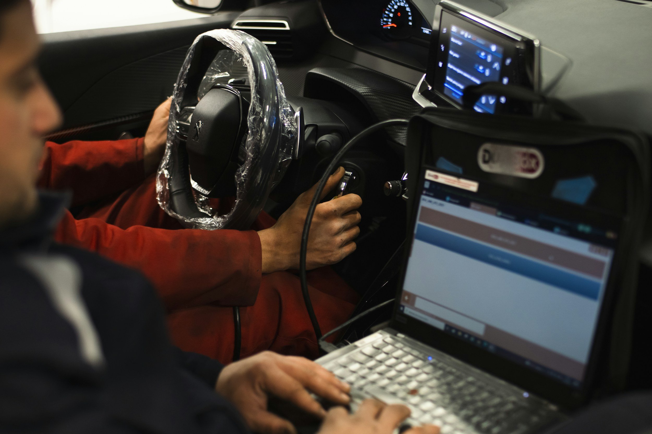 Inside a vehicle with two people: one person seated in the driver's seat, wearing a red jacket, testing or working on the steering wheel covered with plastic, and the other person in the passenger seat using a laptop. The dashboard and two screens are visible inside the car.