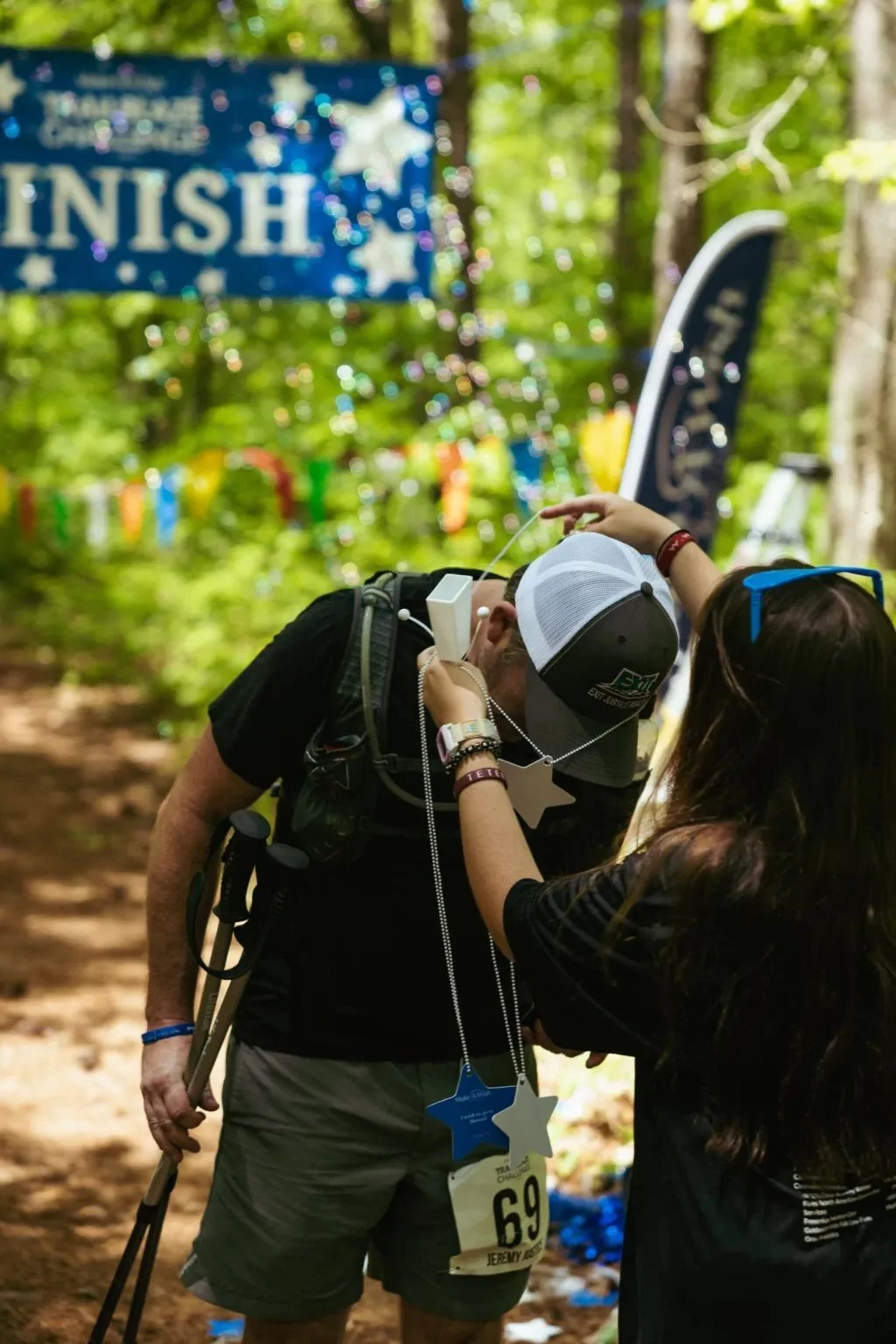 A man at the finish line of a trail race receiving a medal from a woman in a wooded outdoor setting with a decorated background.