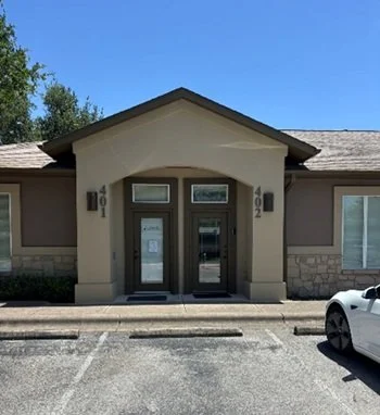 Front view of a beige commercial building with double glass doors and the number 402 above the entrance, parked car on the right, clear blue sky overhead.