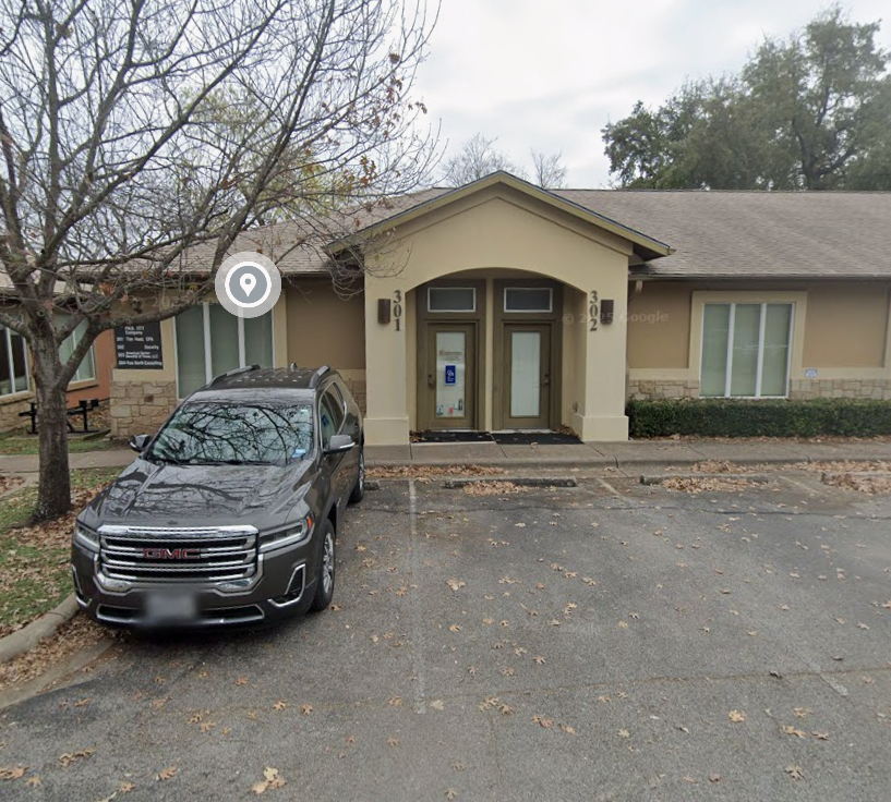 A beige single-story building with an arched entryway numbered 301 and 302, two glass-paneled doors, a tree on the left, and a GMC SUV parked in front on an asphalt lot scattered with fallen leaves.