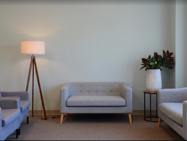 Living room with a beige sofa, gray armchair, floor lamp, and a tall white vase with green and red plants on a black stand, against a light green wall.