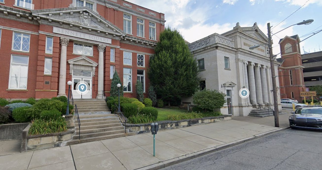 Street view of historic-style buildings, one made of red brick with columns and the other of white stone with columns, parked cars along the curb, street lamps, and a parking meter.