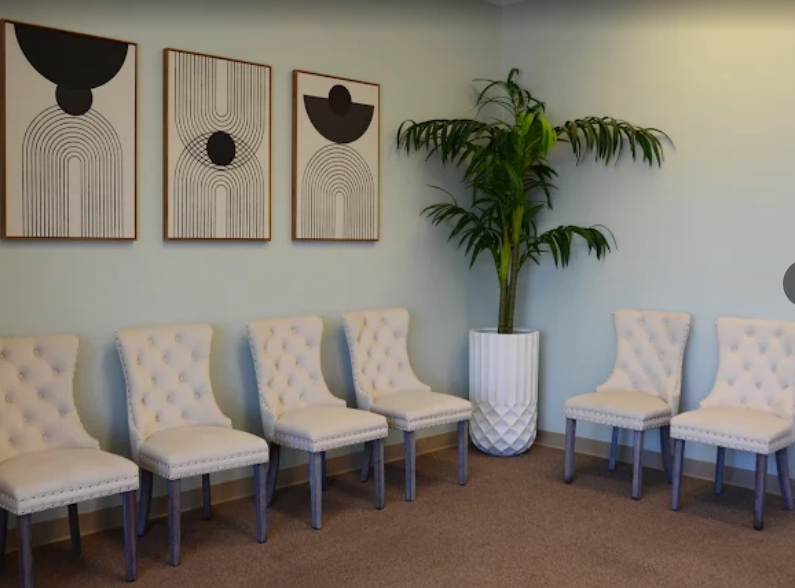 A waiting room with six white velvet tufted chairs arranged along a light-colored wall, decorated with three framed abstract art pieces. A large potted plant is positioned in the corner.