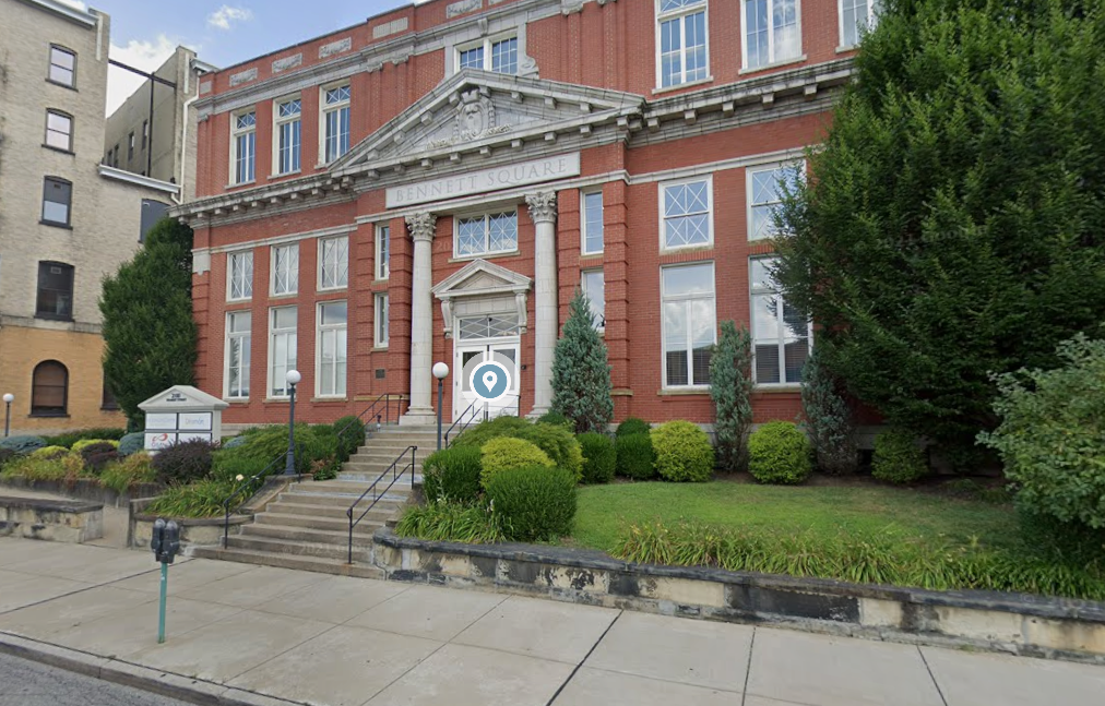 A red brick building labeled Bennett Square with a set of stairs leading up to the entrance, surrounded by landscaped bushes and trees, with a sidewalk and street in the foreground.