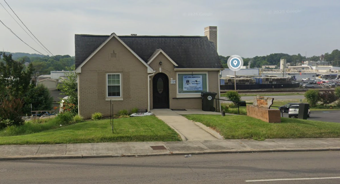 A small beige brick house with a dark gray roof and a chimney, with a sidewalk leading to the front door, a window displaying a sign, and a lawn with some plants and benches outside. There are commercial buildings and parking lot in the background.