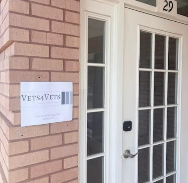 A brick house entrance with a white door featuring glass panels and a keypad lock. There is a sign on the brick wall that reads 'VETS4VETS' with additional text below.