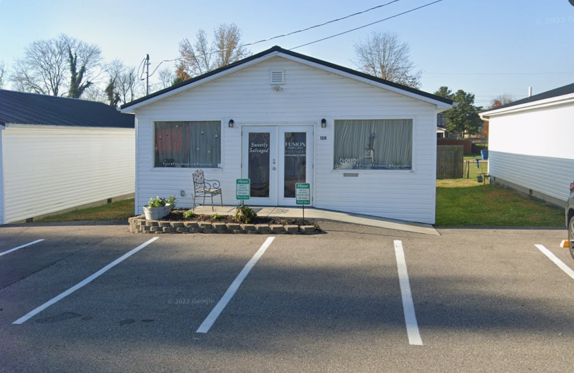 Front view of a small white commercial building with parking spaces in front, two signs in the parking lot, a porch with a chair and potted plant, and windows with curtains.