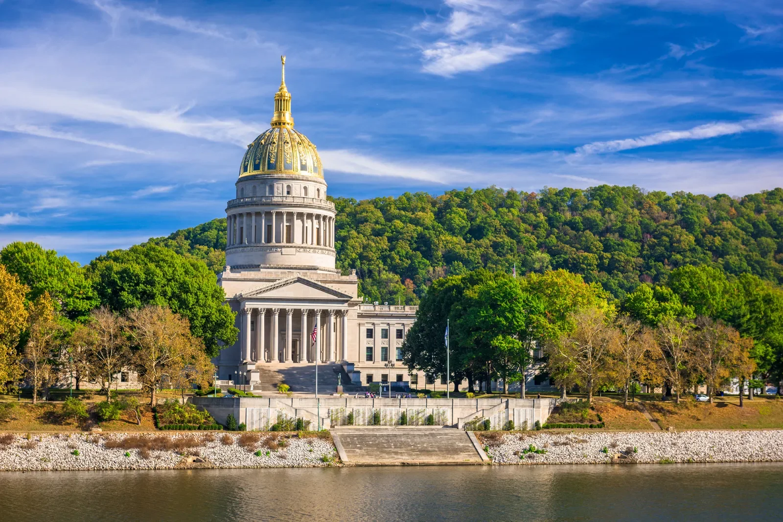 The Arkansas State Capitol building featuring a gold dome with a statue on top, set against a backdrop of a green forested hill and a partly cloudy sky. The building is near a body of water with stairs leading up to the entrance, and surrounded by trees.