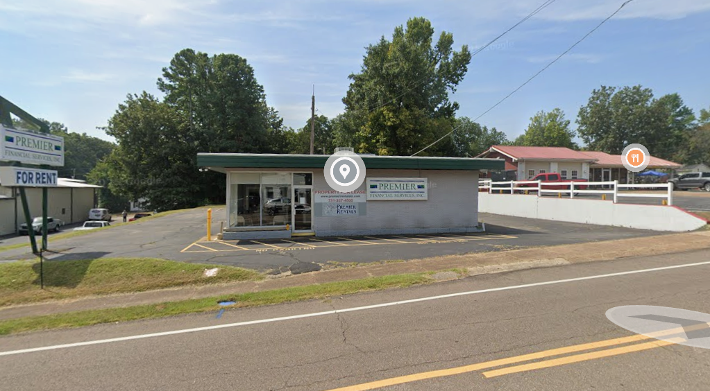 Small commercial building with a sign for Premier Financial and Rent, located on a roadside with parking spaces in front, surrounded by trees and adjacent to another building.
