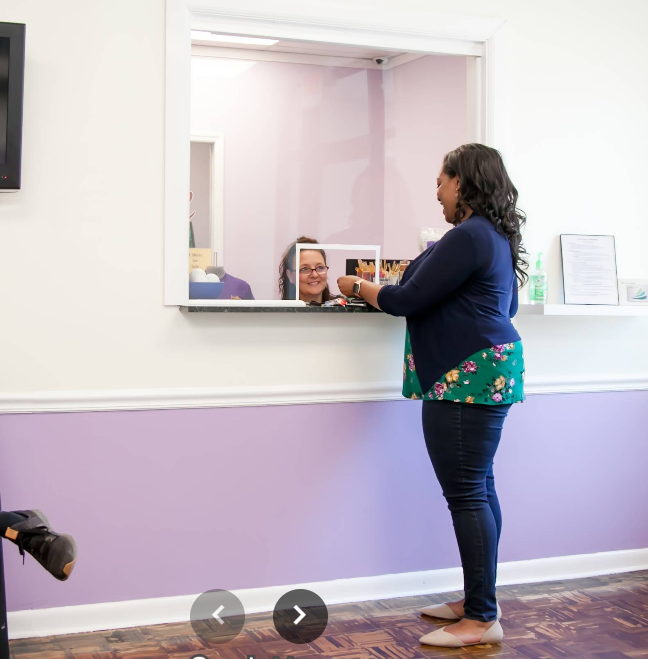 Woman at a reception desk interacting with a client through a window, smiling, with a computer monitor and framed photos on the counter.
