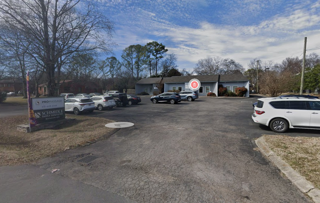 A parking lot with several parked cars in front of a one-story blue building, and a sign on the left that reads 'Schmidle Family Dentistry' and 'Pro Massage', with trees and clouds in the background.