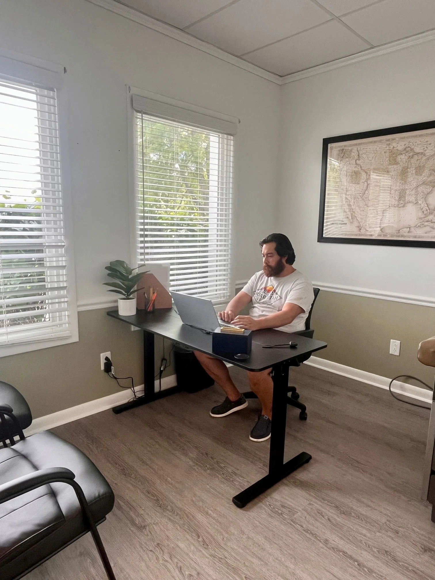A man sitting at a black desk on a black office chair working on a silver laptop in a bright room with white walls, large windows with white blinds, a framed map on the wall, and a gray sofa in the foreground.