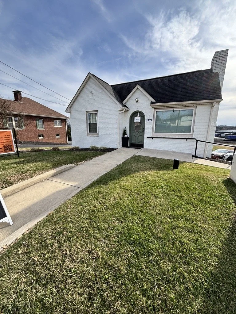 A small white brick house with a dark gray roof and a chimney, with a sidewalk leading to the front door, a window displaying a sign, and a lawn with some plants and benches outside. There are commercial buildings and parking lot in the background.