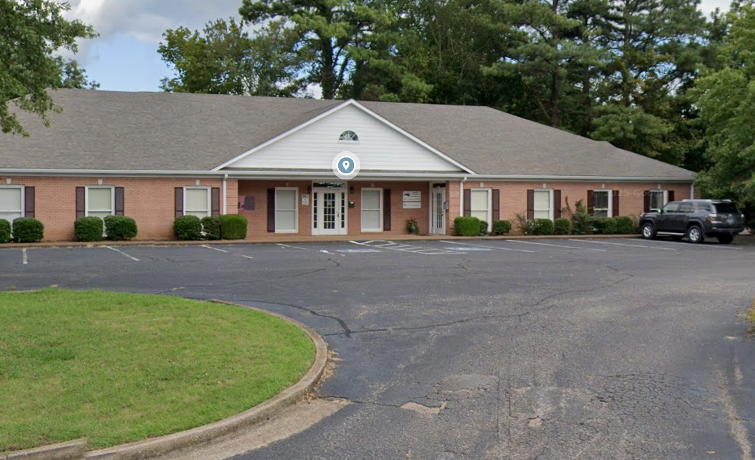 A single-story brick building with white siding and a gabled roof, surrounded by trees, with several parked cars in the parking lot.