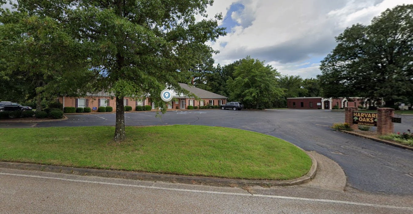 A parking lot with a brick building labeled Harvard Oaks, surrounded by trees and landscaped flower beds, under partly cloudy skies.