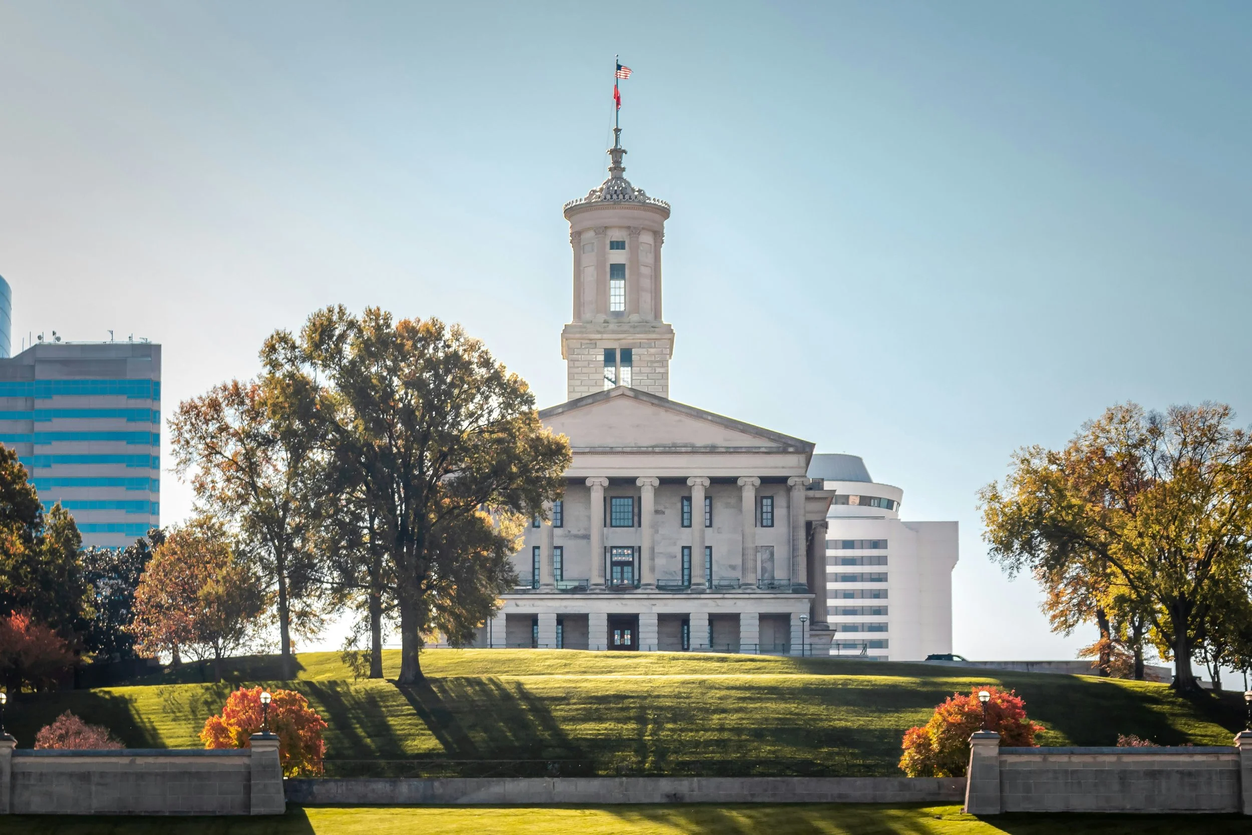 The Ohio Statehouse, a historic government building with columns, atop a grassy hill with trees, with modern high-rise buildings in the background, under a clear blue sky.