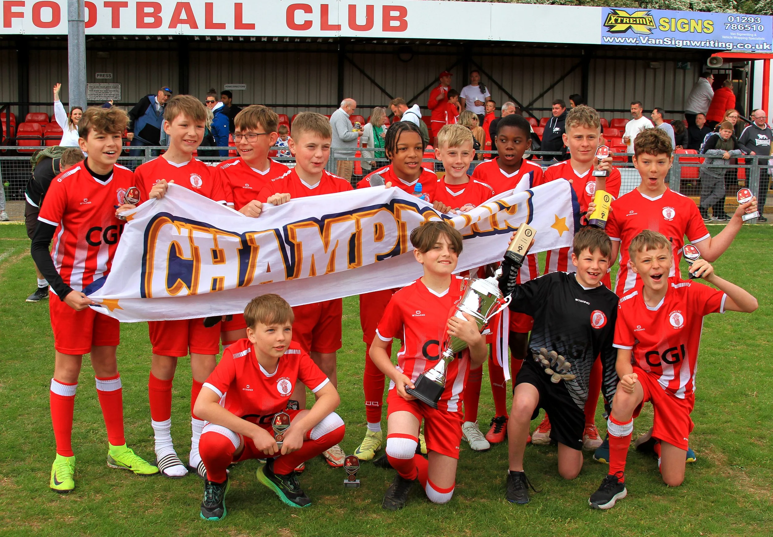 A group of young boys in a red and white kit, celebrating after winning a championship. They are holding a large banner reading 'CHAMPIONS' and some are holding trophies and medals. There are spectators and team staff in the background.