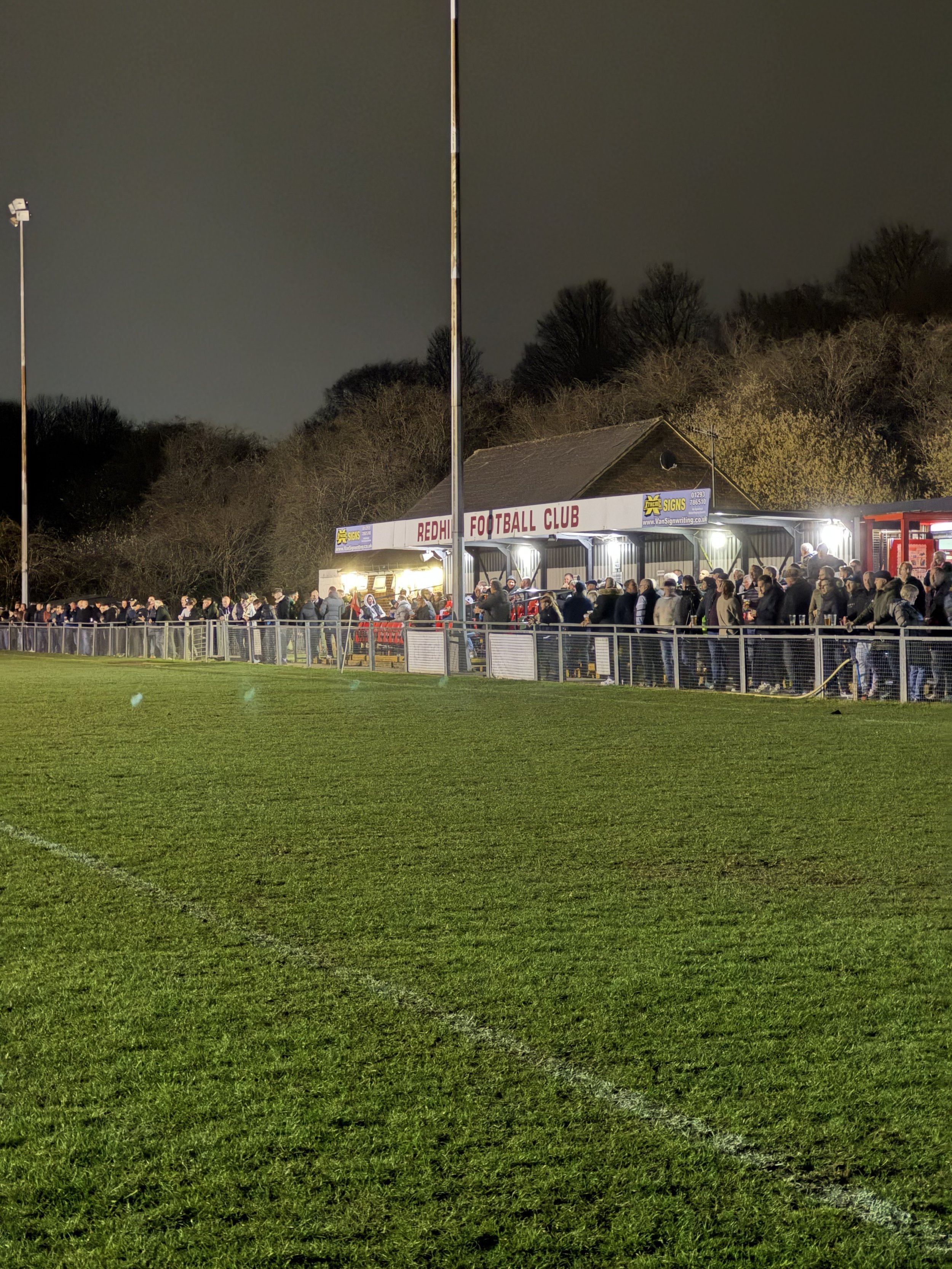 Night scene of a football field with a crowd of spectators standing along the fence and a clubhouse building that reads "Redhill Football Club" in the background.